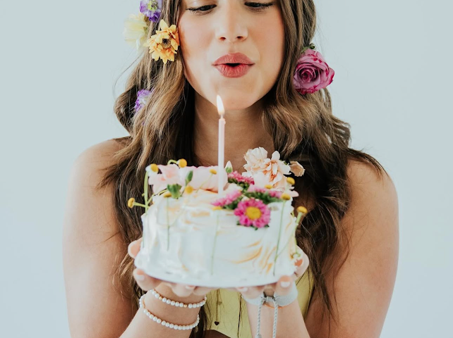 Young woman with floral hair accessories blowing out a candle on a decorated white birthday cake.