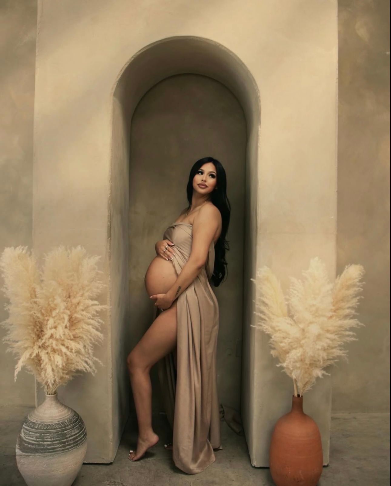 Pregnant woman in beige draped gown poses in arched niche flanked by pampas grass in terracotta vases.