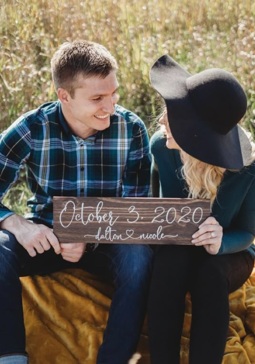 Couple in matching plaid shirts holding a wooden sign displaying their wedding date in a rustic outdoor setting.