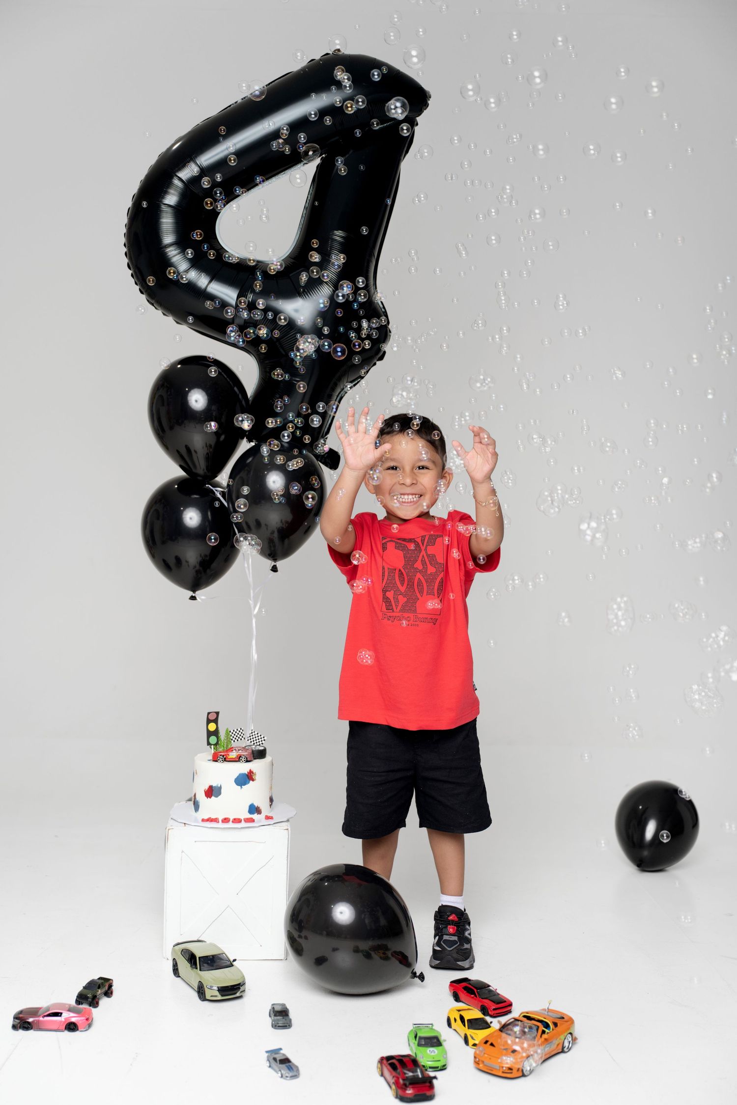 A joyful boy celebrates his 4th birthday with black balloons, toy cars, and a cake while playing with bubbles.