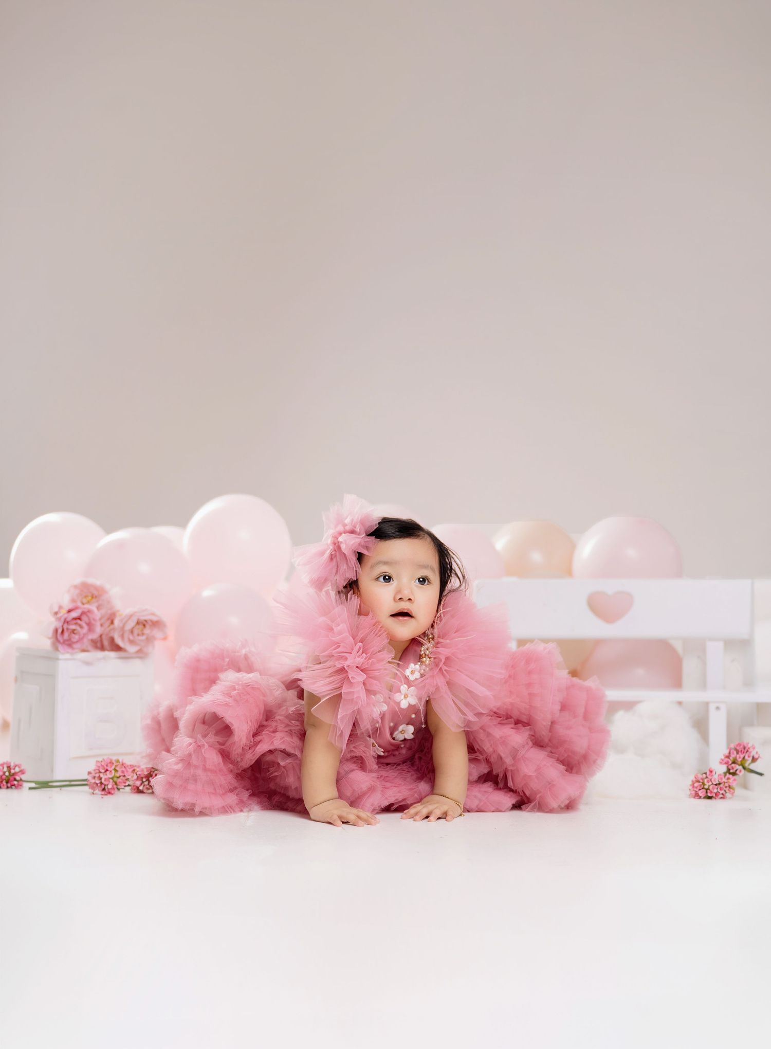 Baby girl in pink tulle dress poses among pink balloons and flowers for a first birthday photo session.