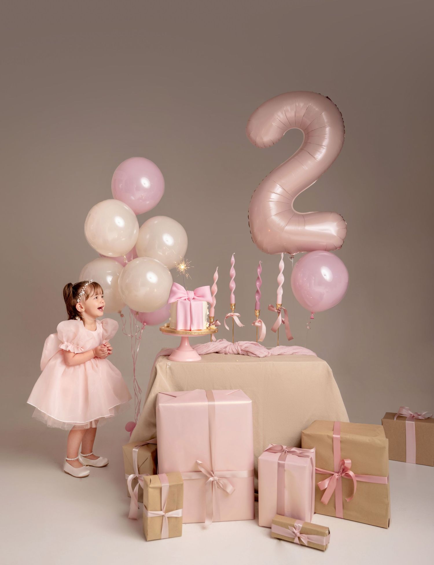 A toddler girl in a pink dress celebrates her 2nd birthday with balloons, gifts, and a sparkly cake.