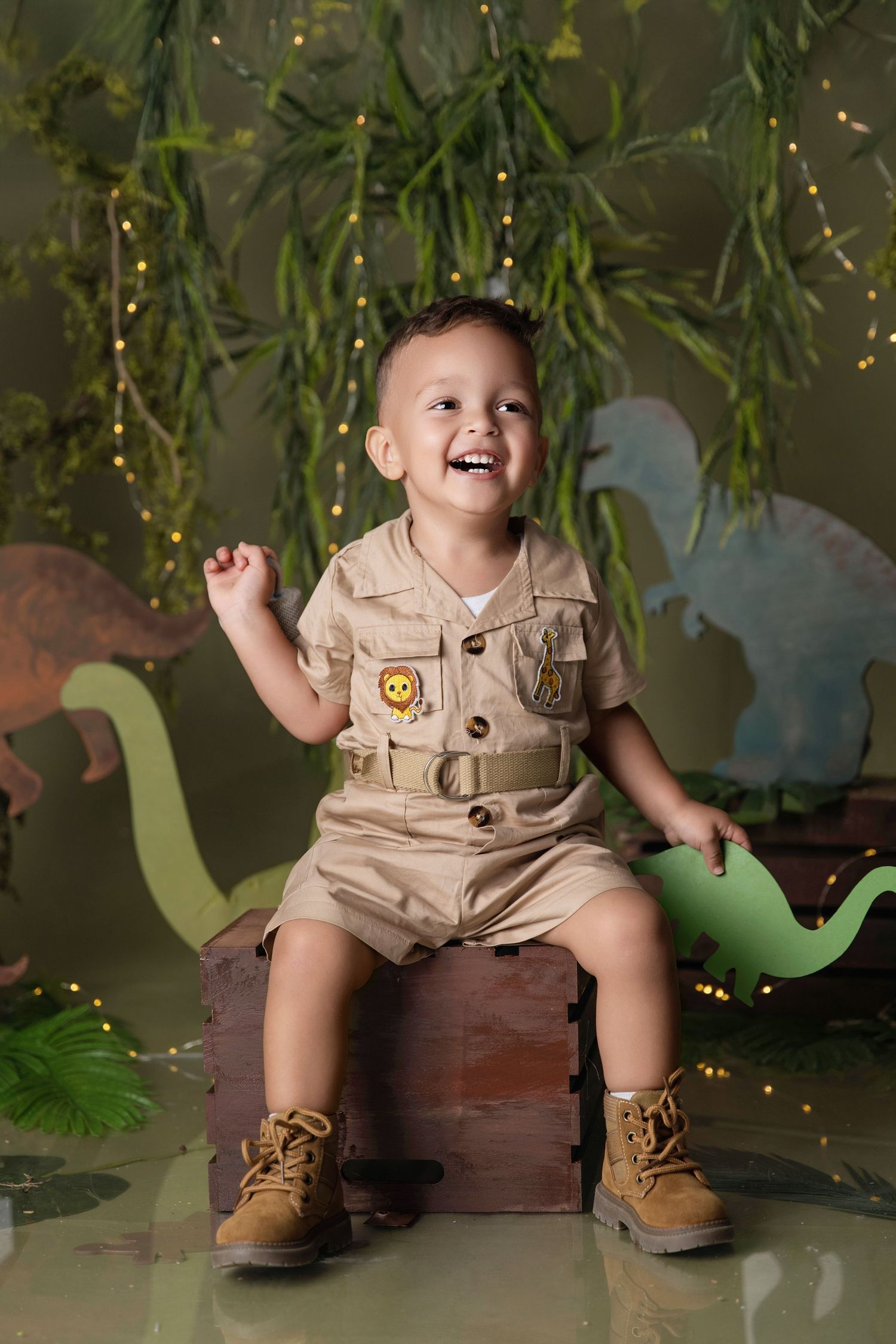 Laughing toddler boy in safari outfit sits on wooden crate with dinosaur decorations and jungle backdrop.