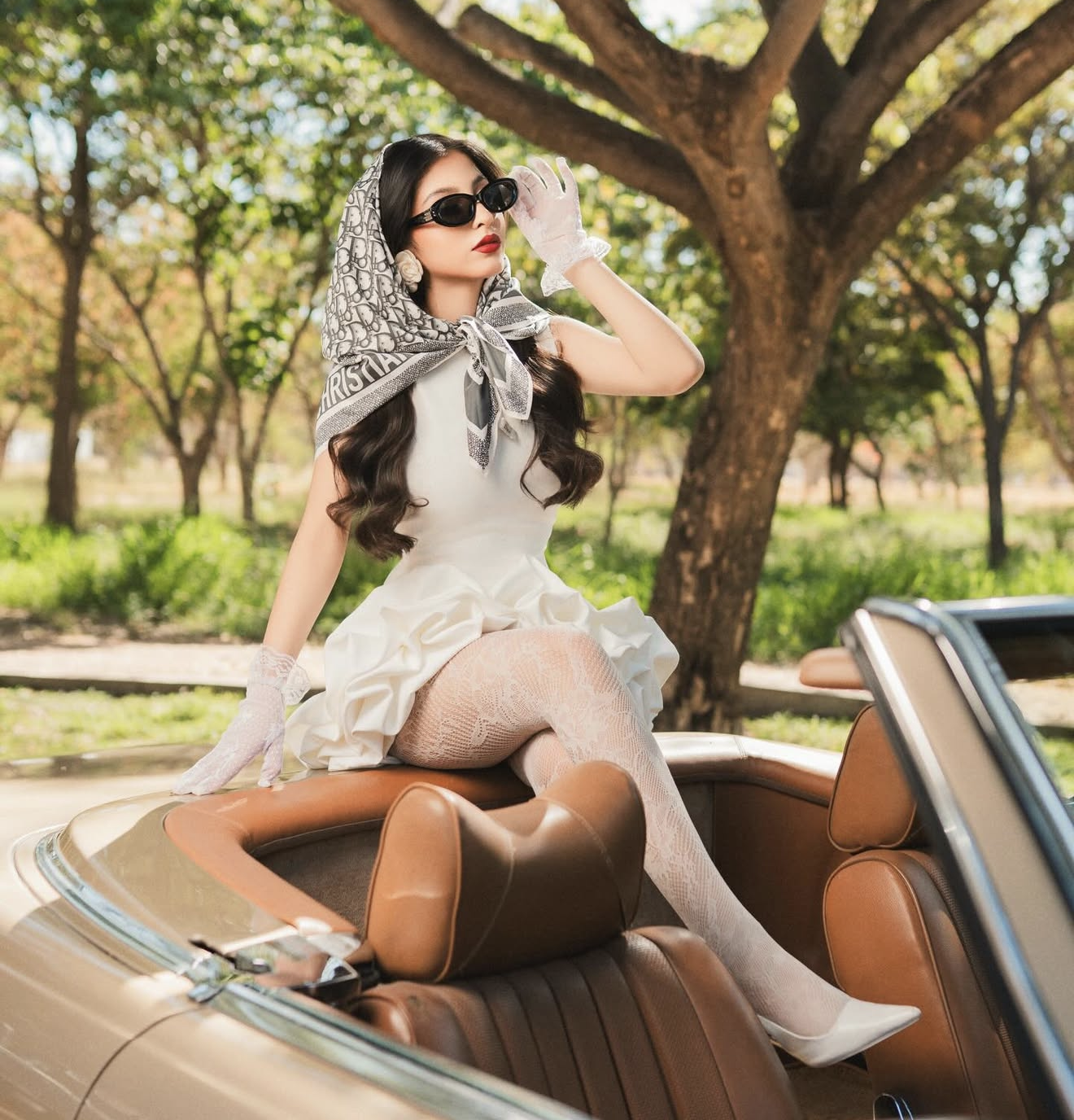 Elegant woman in white dress, head scarf, sunglasses, and gloves posing in a vintage convertible car outdoors.