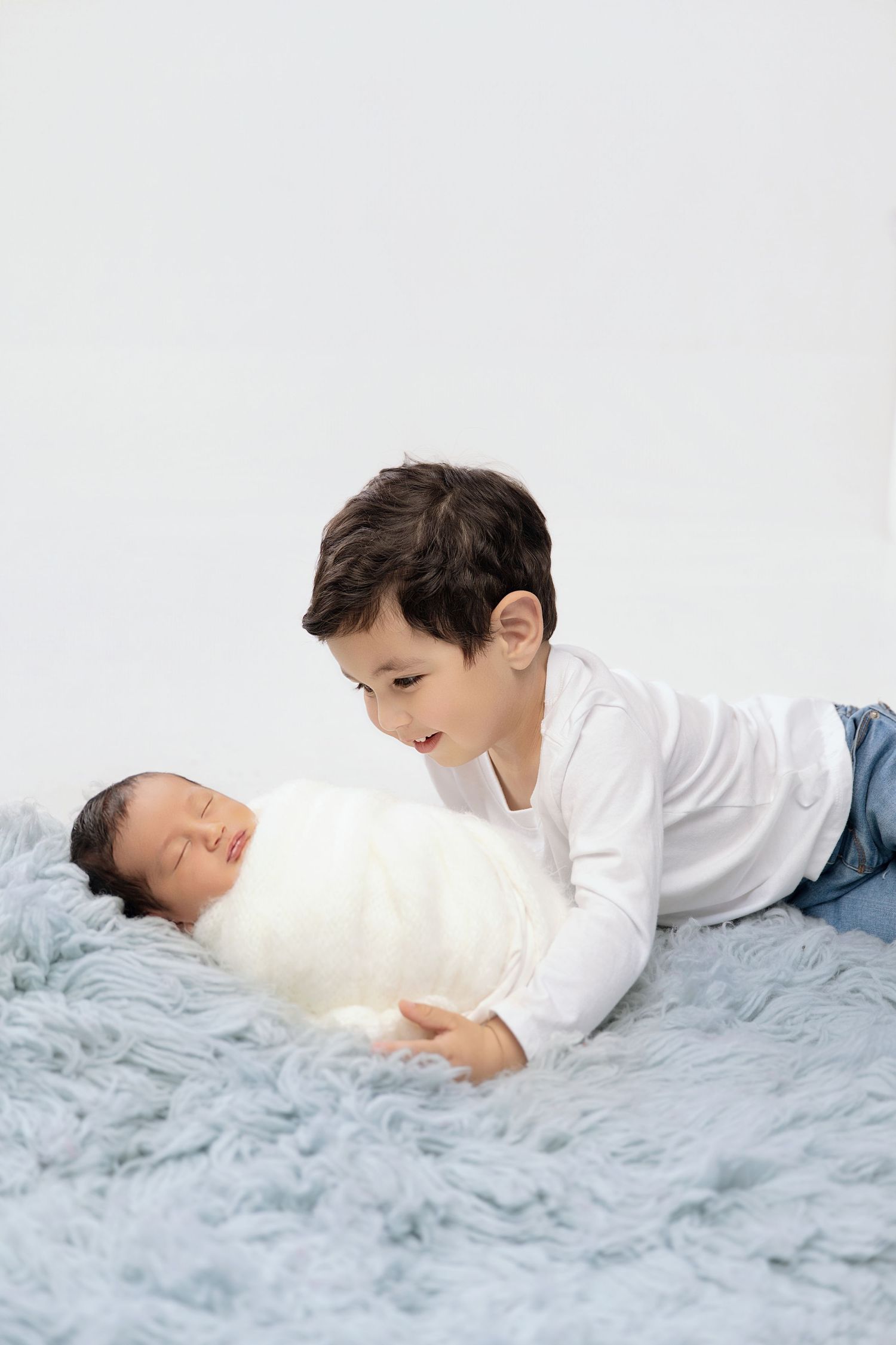 A young boy in a white shirt lies on a blue blanket, smiling at a sleeping newborn baby wrapped in white.