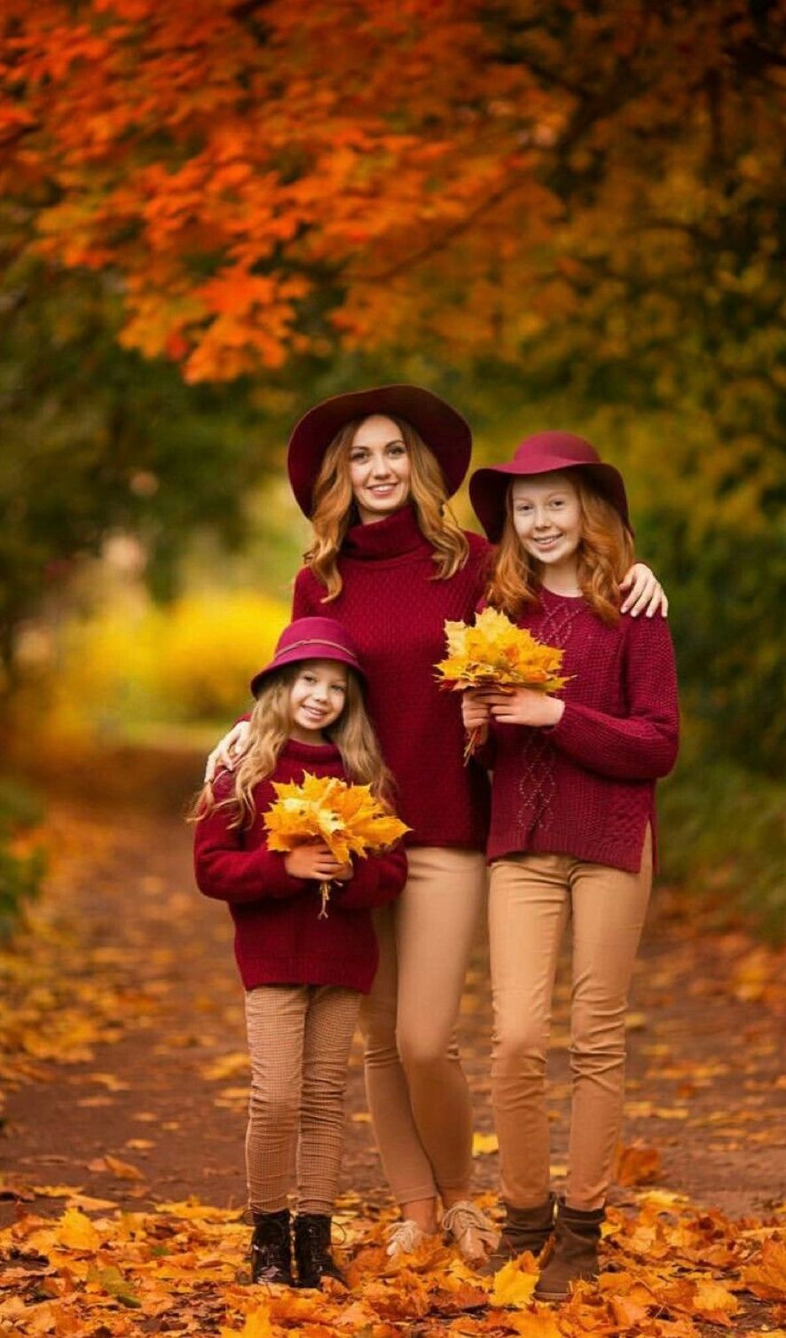 Three people in matching burgundy sweaters and autumn hats pose together on a leaf-covered path amid vibrant fall foliage.