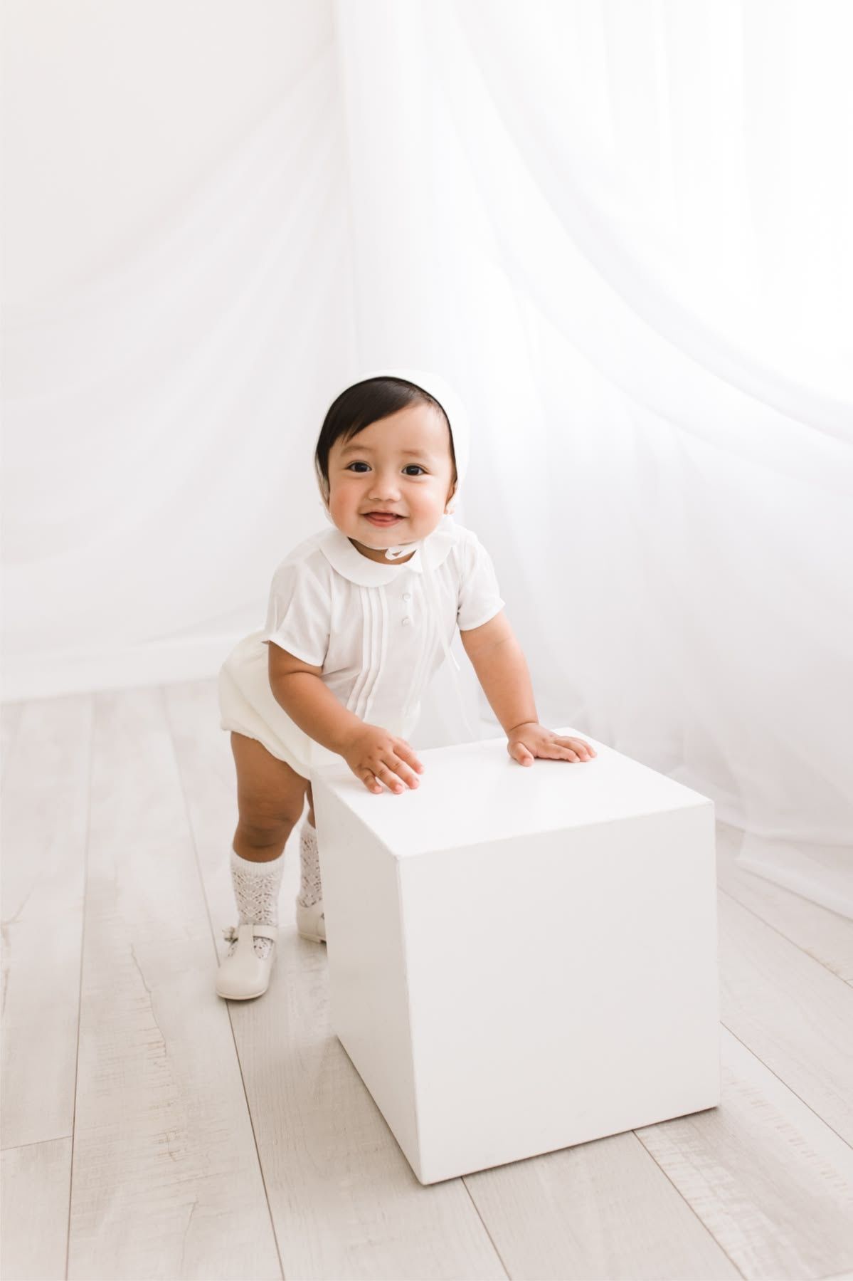 A smiling baby boy in a white romper and knee socks stands holding a white cube against a white backdrop.