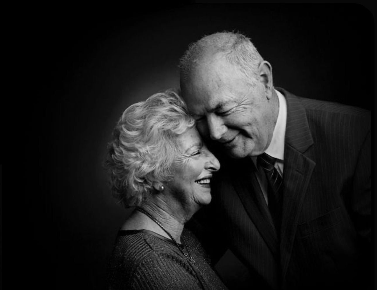 Black and white portrait of a smiling elderly couple sharing a tender moment, touching foreheads affectionately.