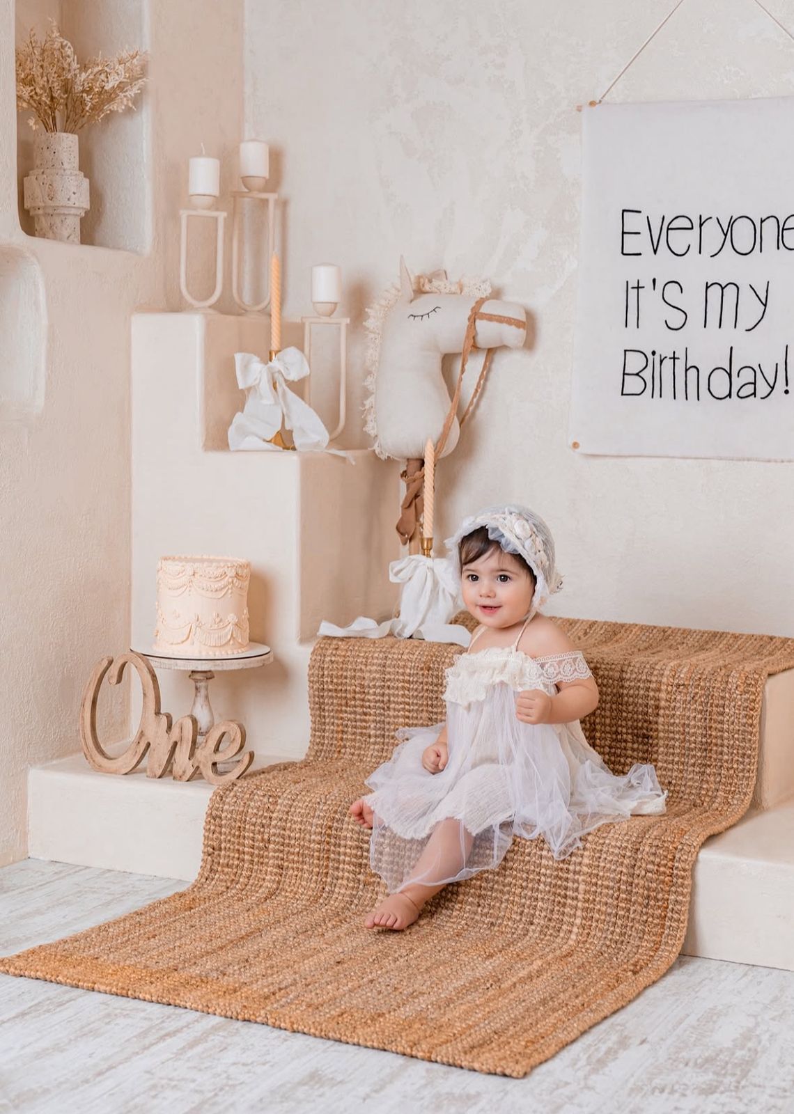 A baby in a white dress sits on a textured mat for a first birthday photoshoot with decorative signs and props.