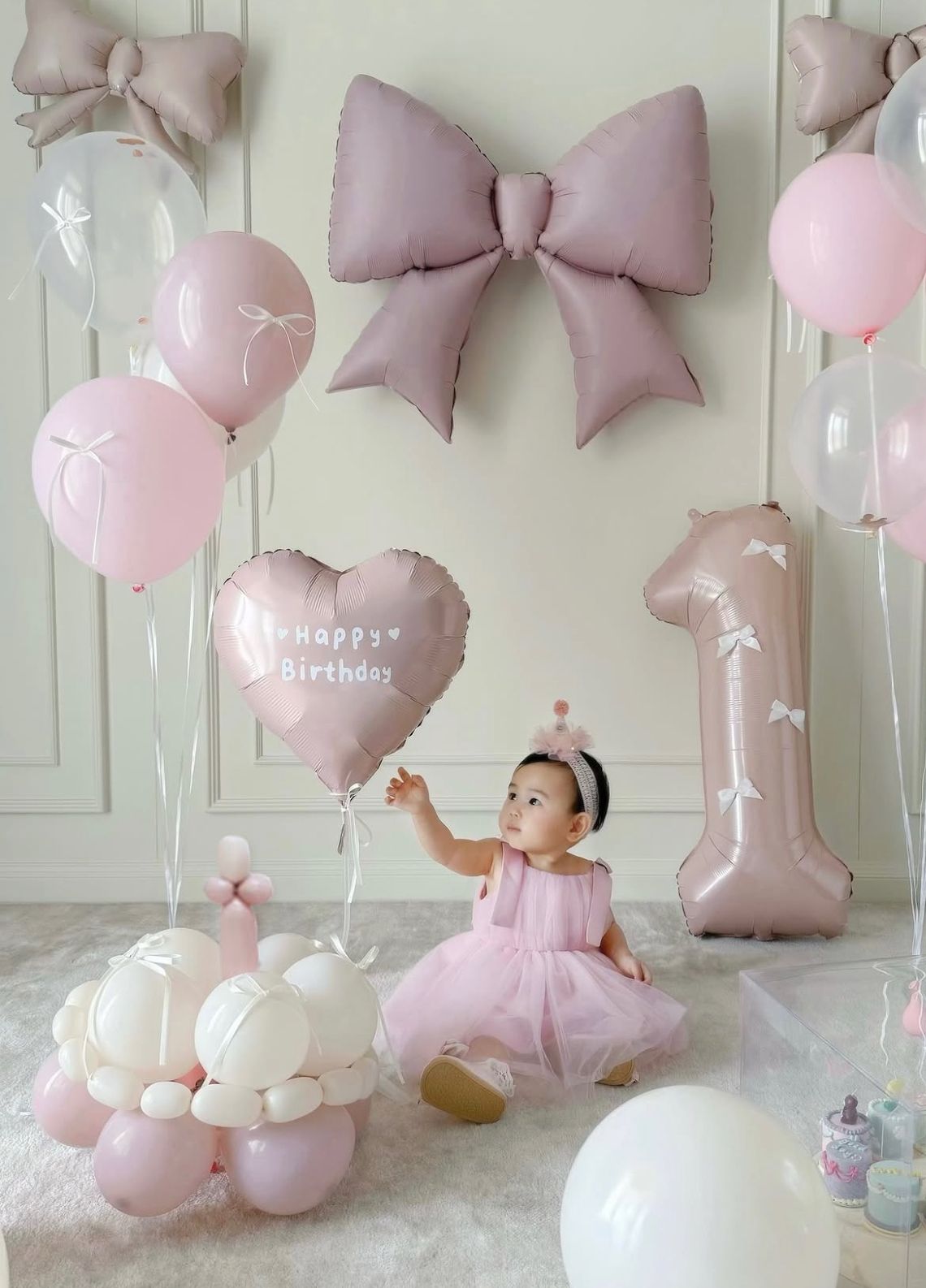 Dreamy first birthday photo setup with pink and mauve bows, balloons, and tulle decorations against a white wall.