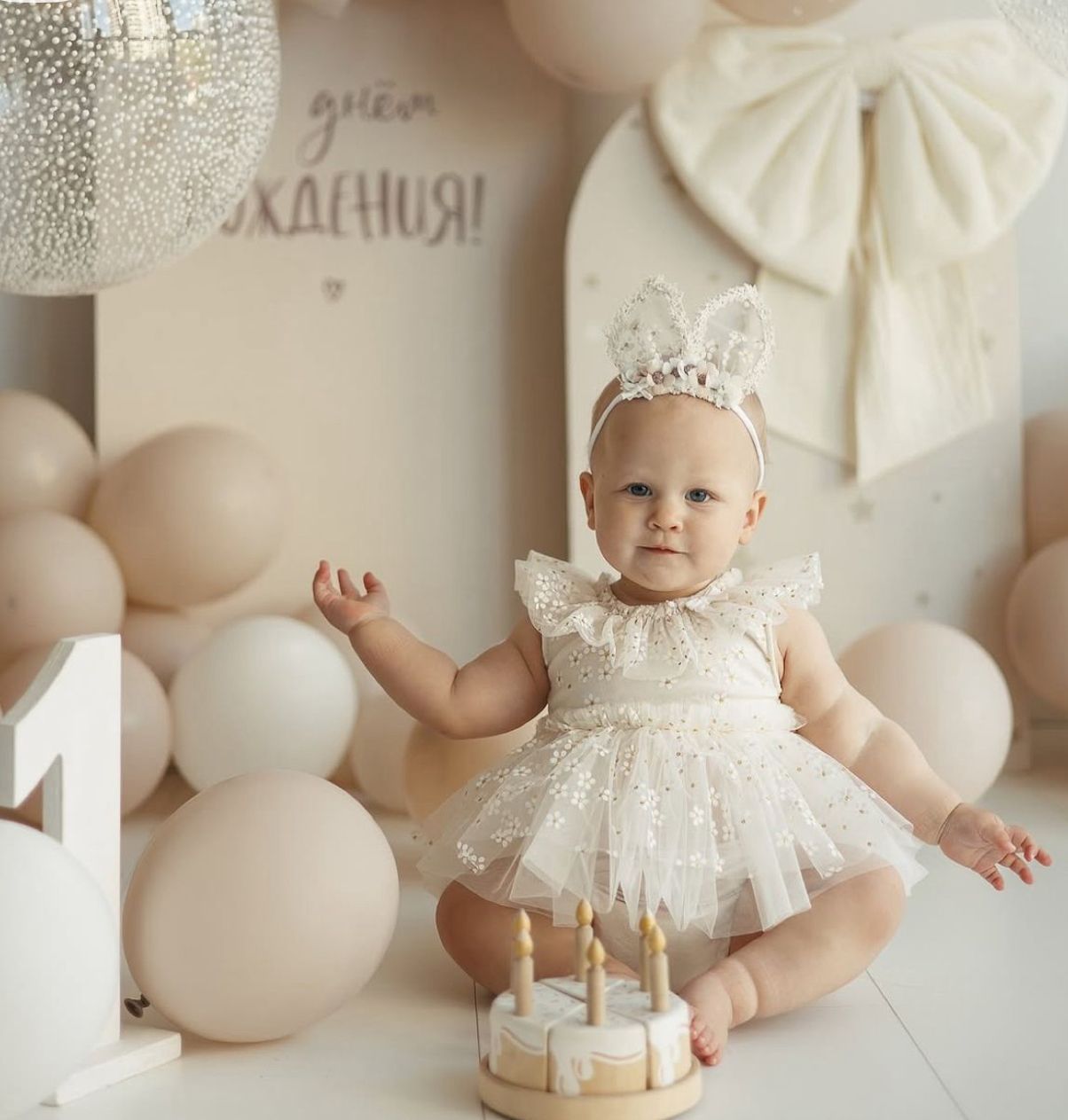 A baby in a white lace dress sits on a cream backdrop surrounded by white balloons for a first birthday photoshoot.