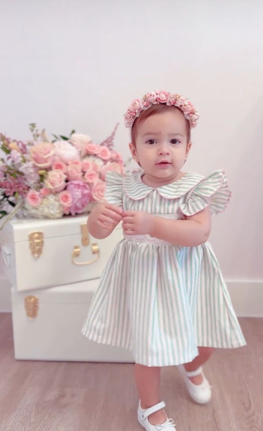 Baby in striped dress with pink floral crown poses against soft pink backdrop with flowers and white furniture.