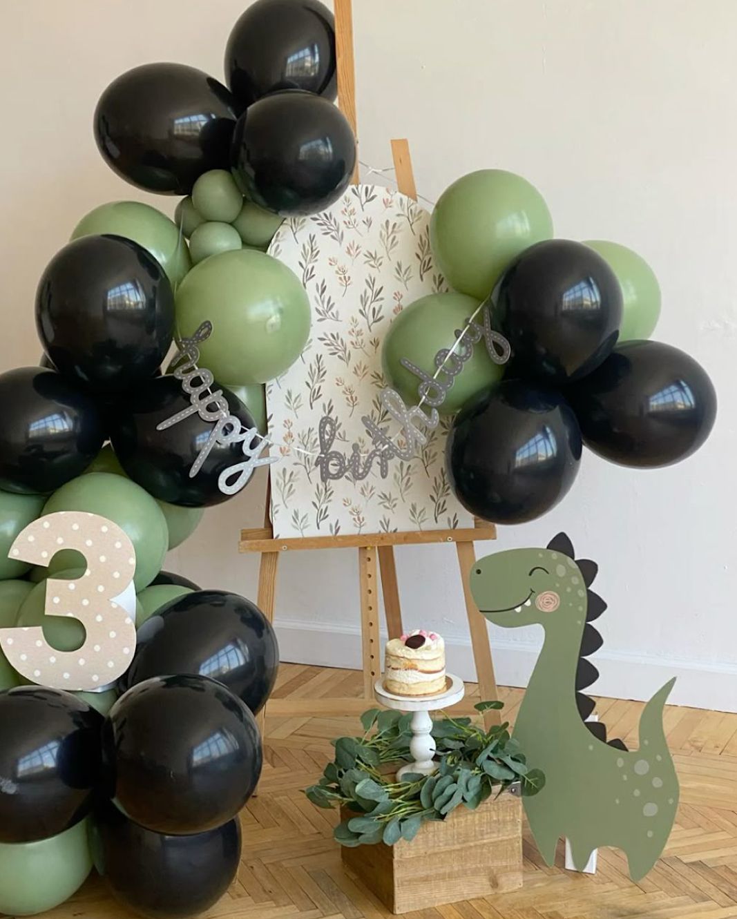 Black and sage green balloon arch with dinosaur theme decorations against a white wall.