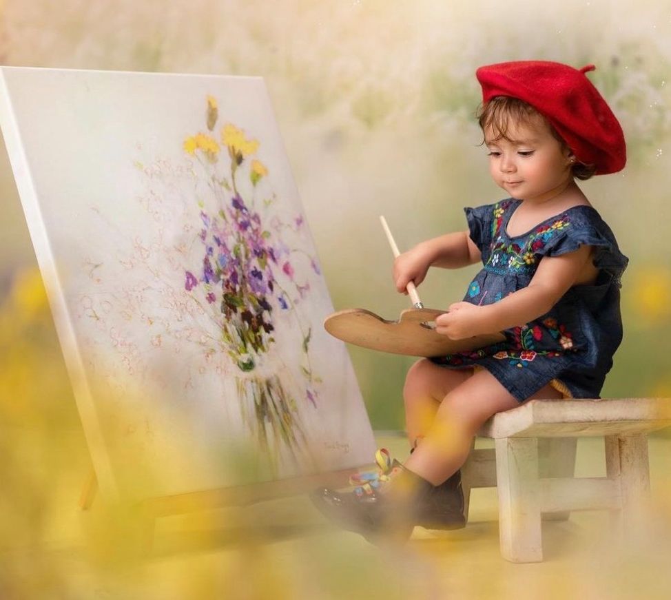 A young artist in a red beret and denim dress sits on a small bench while painting colorful flowers on canvas.