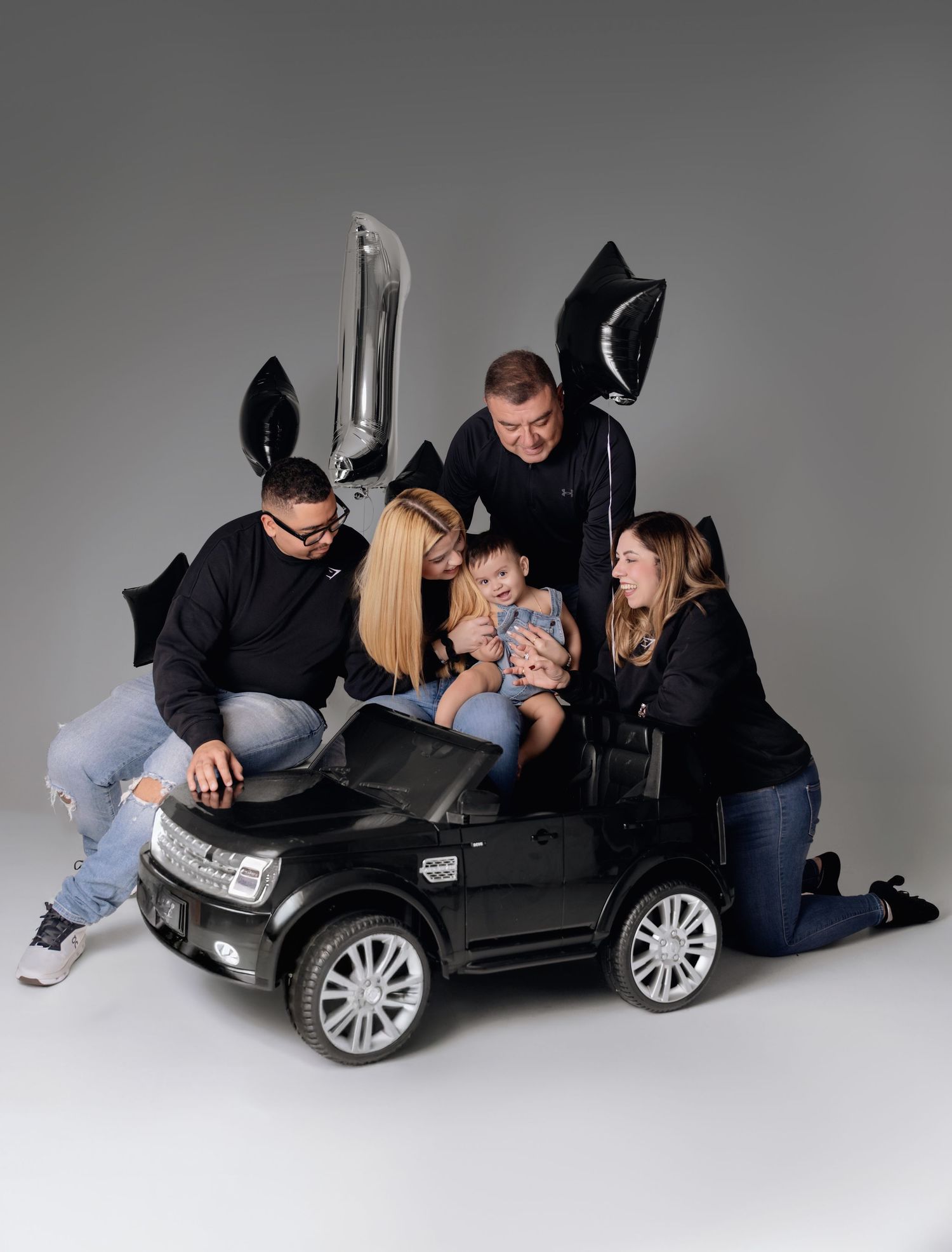 Family celebrating a toddler's birthday around a black toy Land Rover with silver and black balloons.