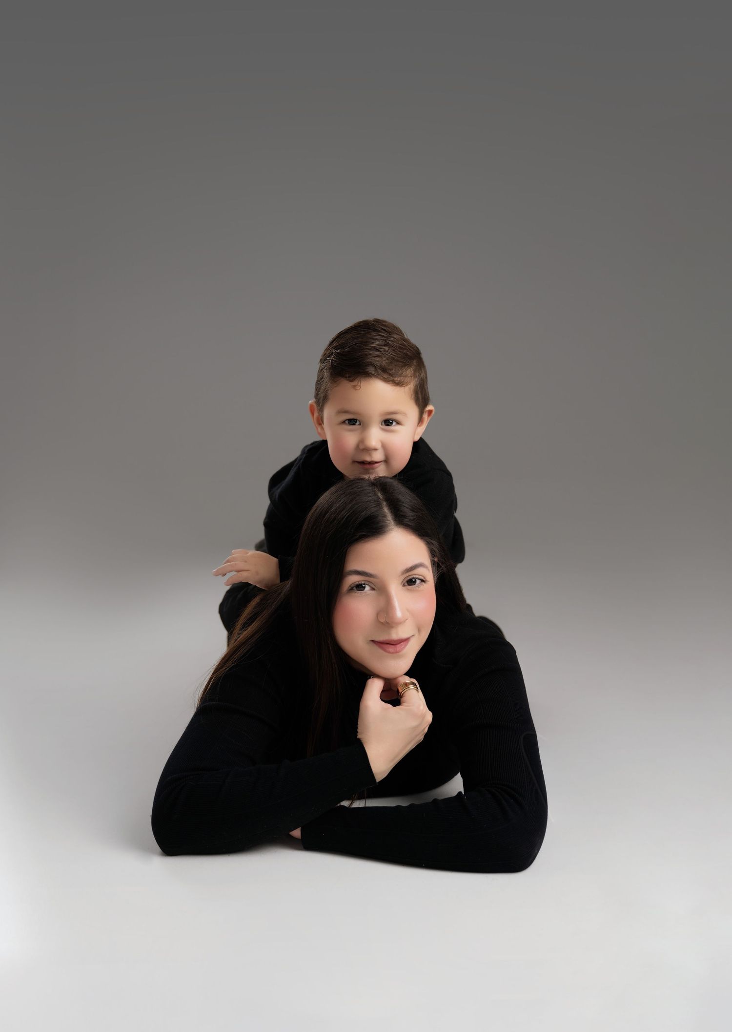 A woman in black lies on the floor while a young boy rests on her back, both smiling at the camera.