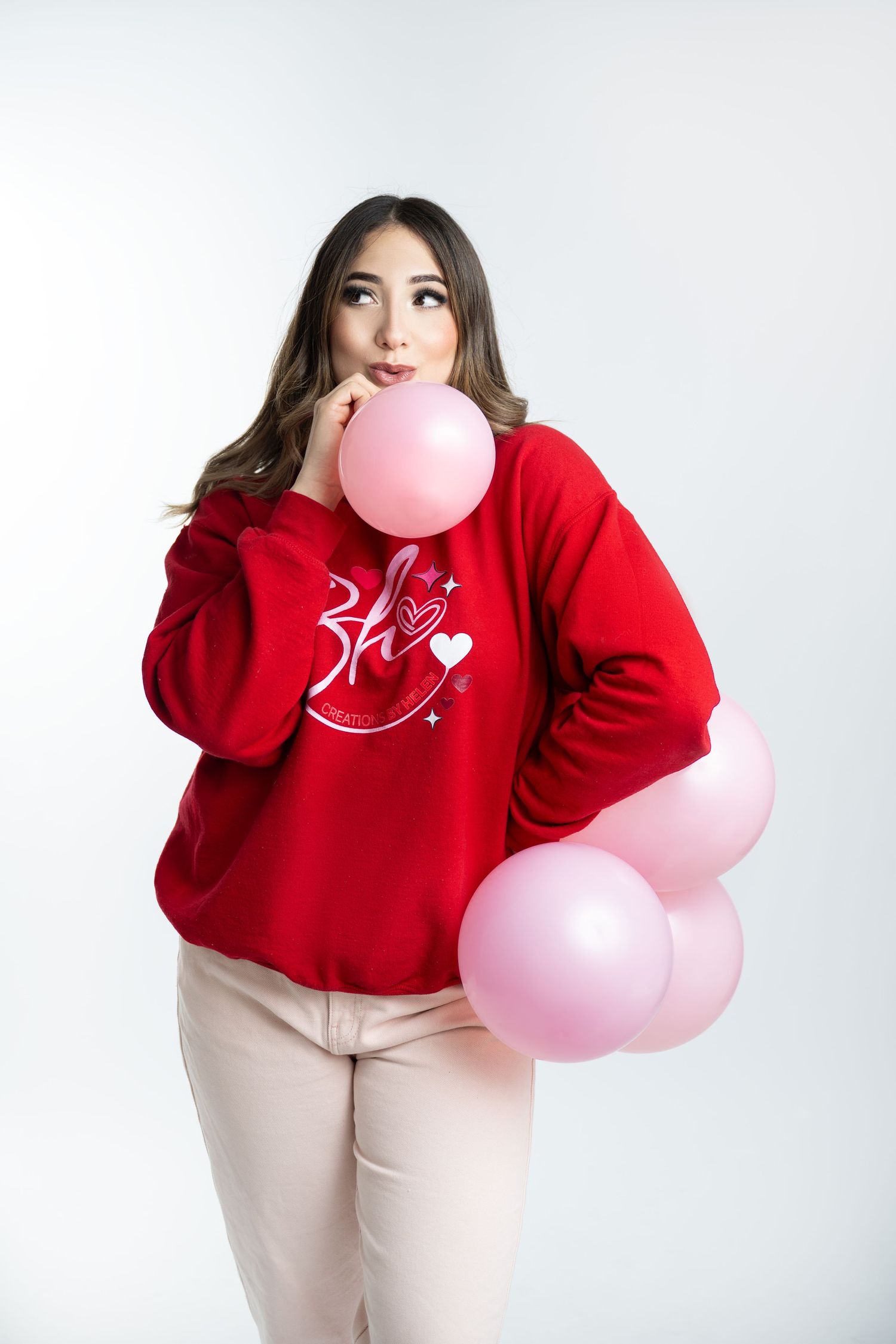 Young woman in red sweatshirt holding pink balloons against white background, posing thoughtfully.