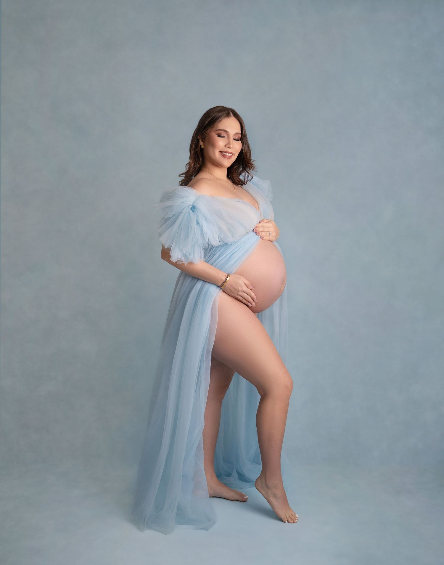 Pregnant woman in sheer light blue tulle maternity gown posing gracefully in a professional studio photo shoot.