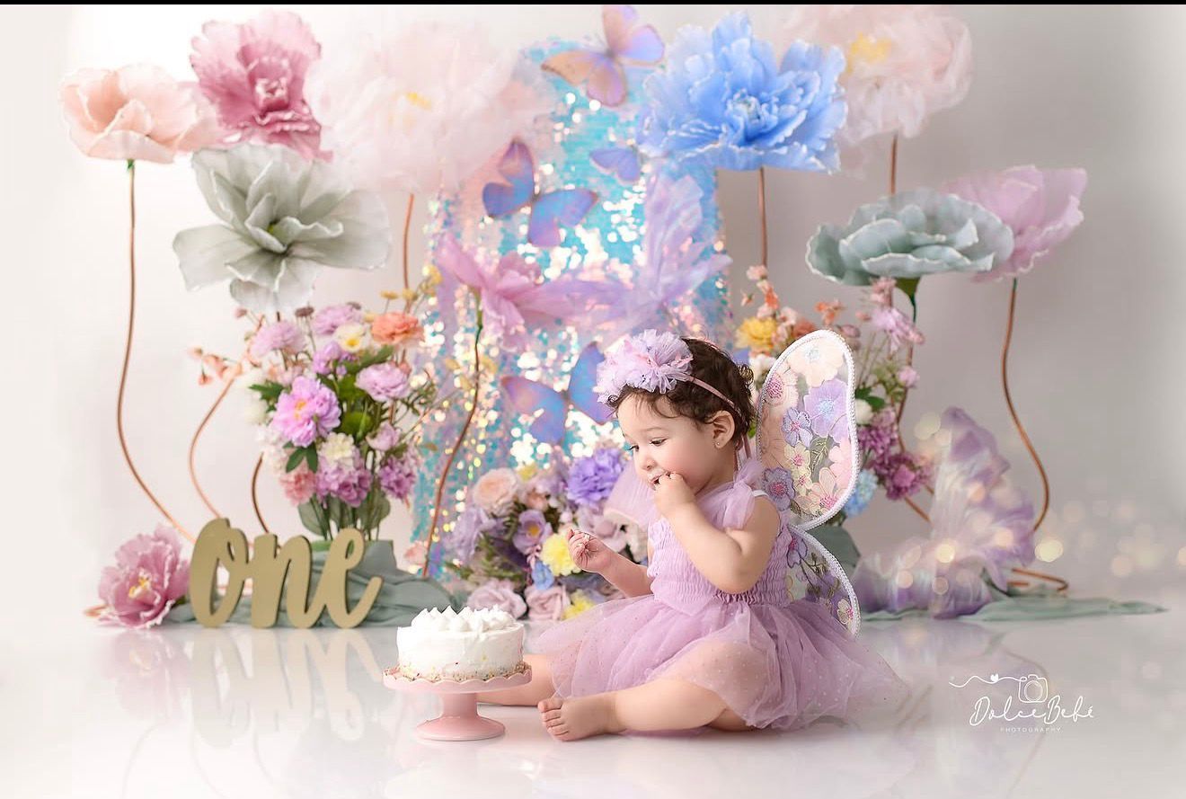 Baby girl in purple tutu with fairy wings celebrates first birthday with cake amid colorful floral decorations.