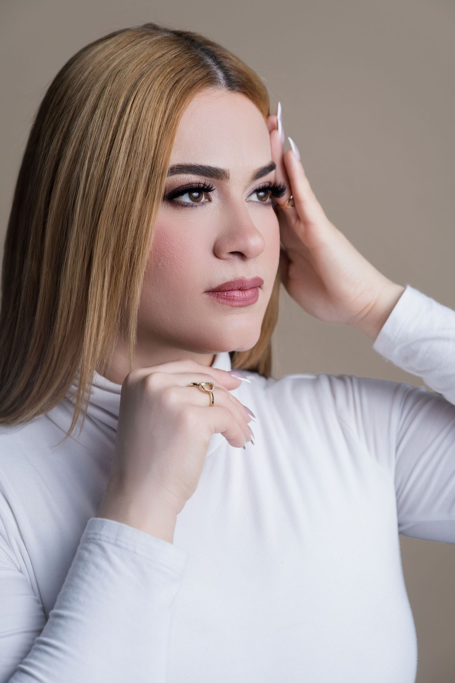 A young woman with blonde hair wearing a white turtleneck top poses thoughtfully with hands near her face, wearing a gold ring.