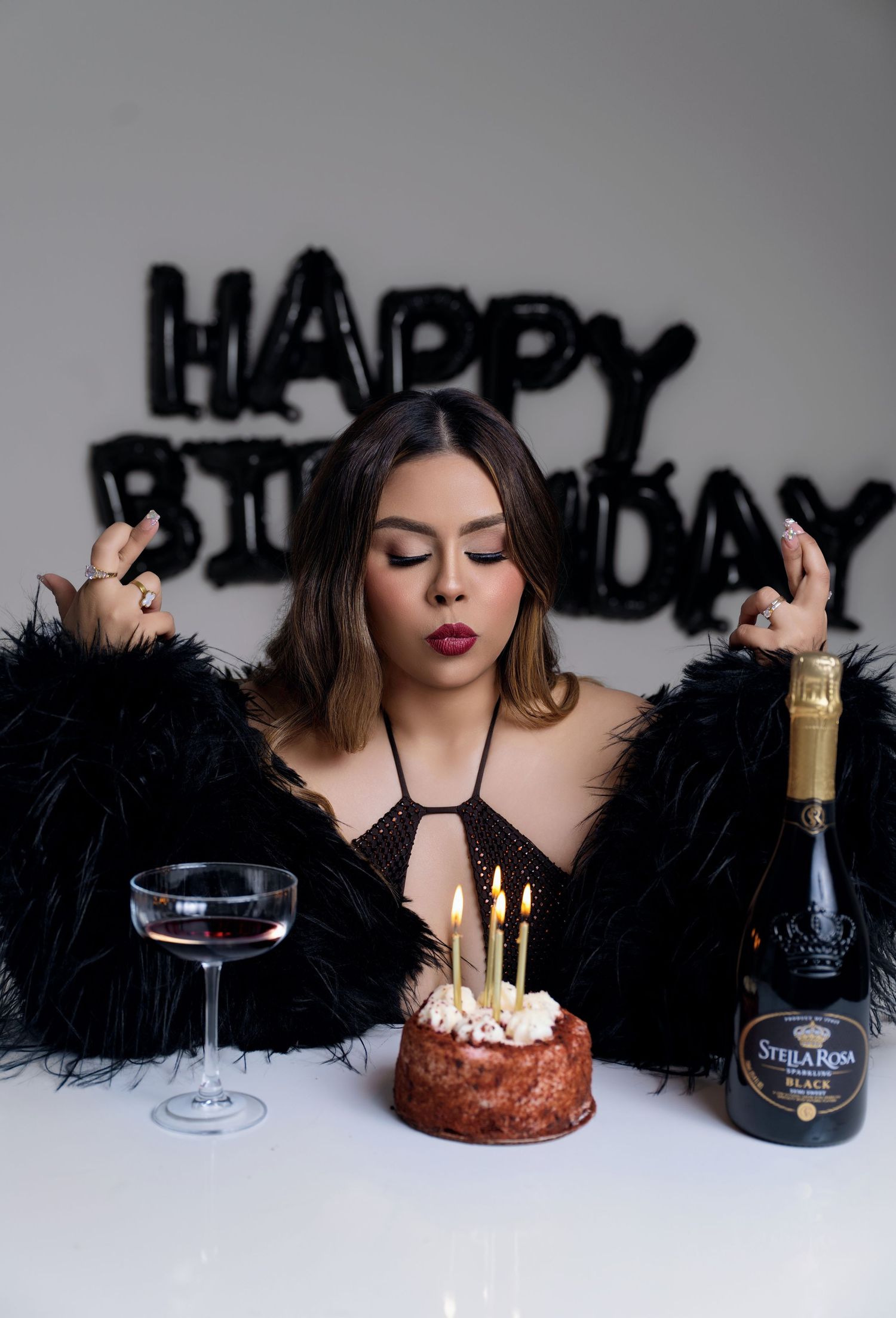 Woman in a black feather jacket poses with a lit birthday cake, champagne coupe and Stella Rosa bottle under black Happy Birthday balloons.