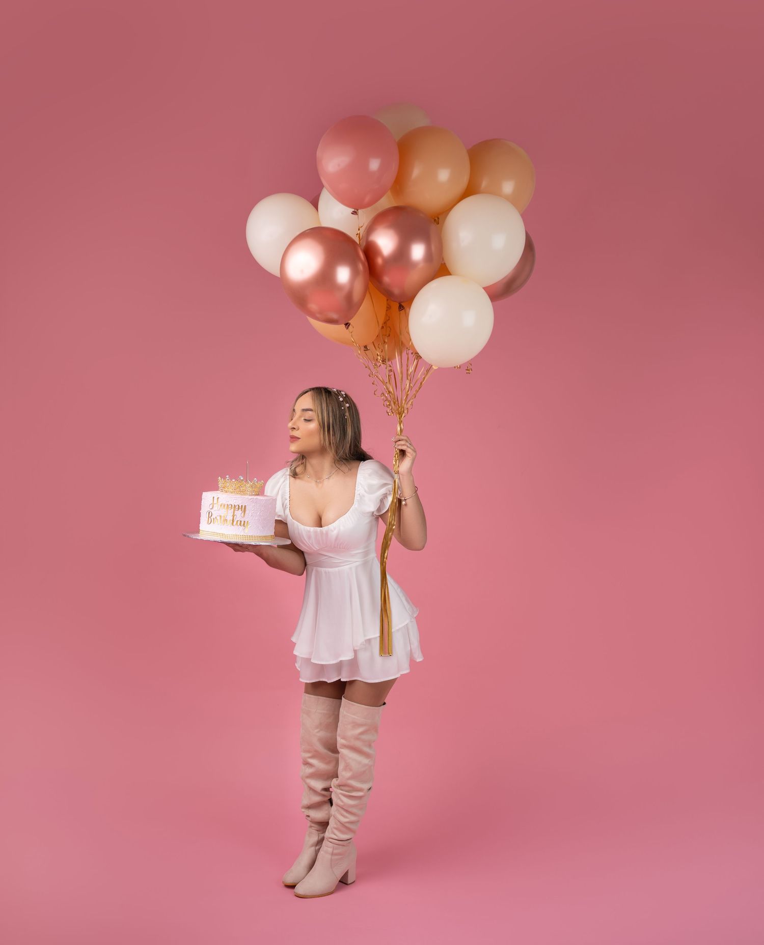 Woman in a white dress and tall blush boots holds a birthday cake and pink, gold and white balloons against a pink studio backdrop.