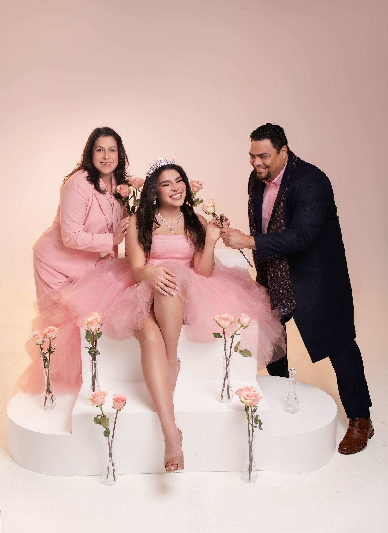 A young woman in a pink tutu and tiara poses with her smiling parents during a quinceañera photo shoot.