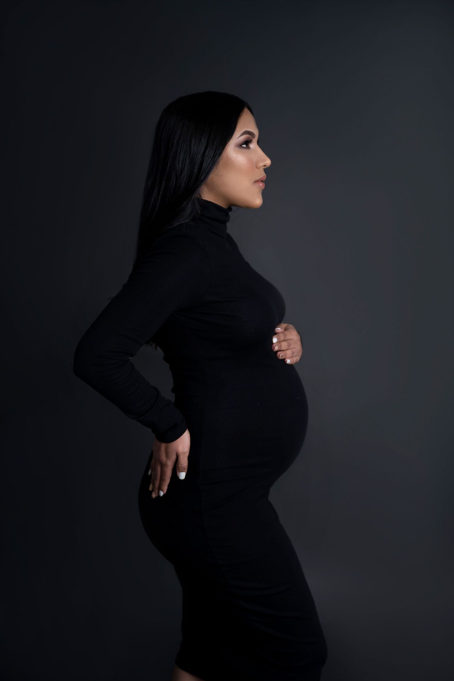 Maternity photo of a silhouette in a black turtleneck dress against dark background.