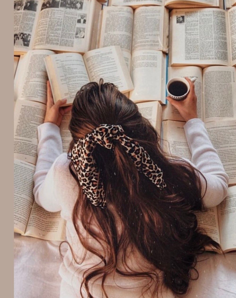 A brunette woman with a leopard print hair scarf reads a book while holding coffee, surrounded by open books.