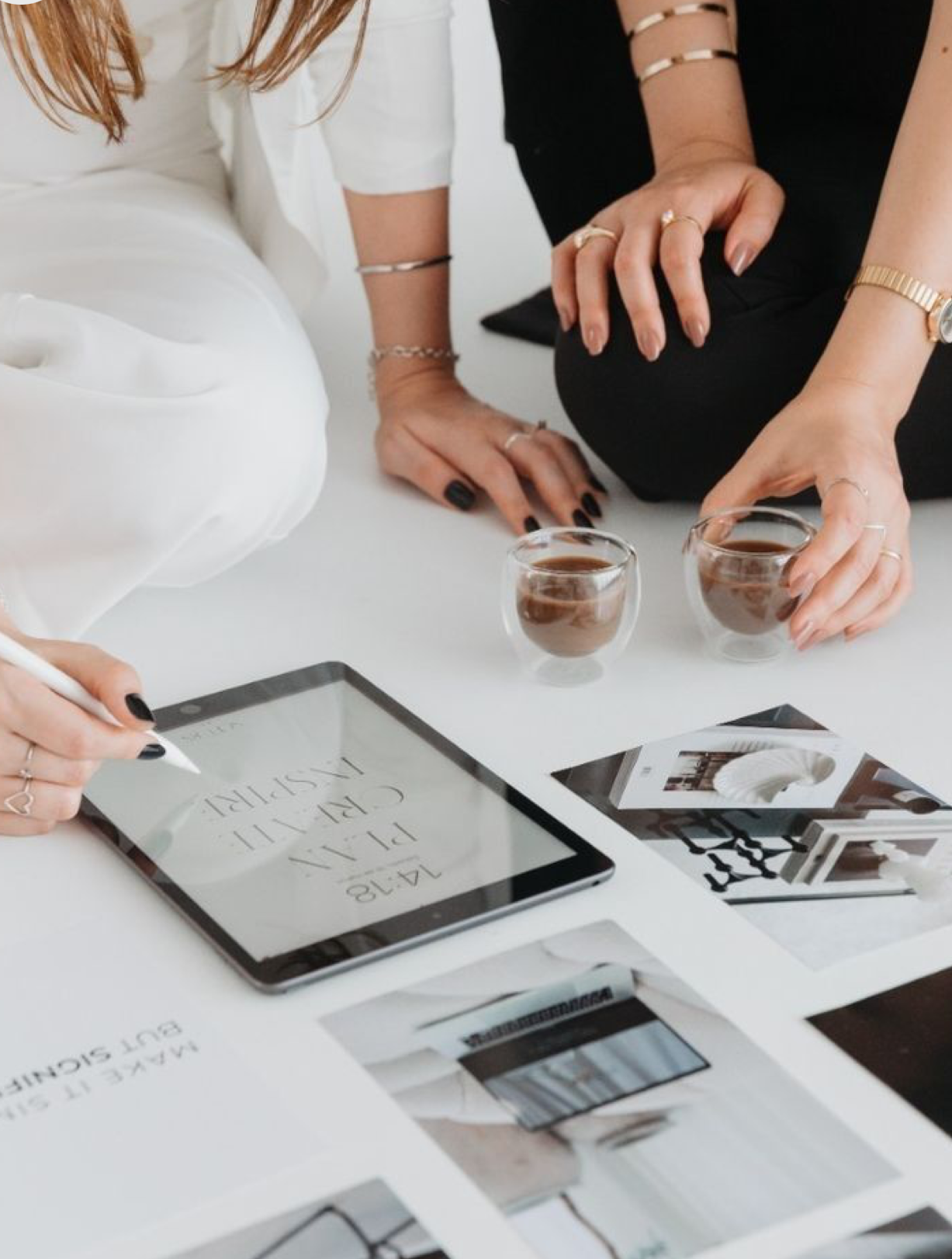 Two women collaborating over interior design mood boards and a tablet on a white table with coffee glasses.