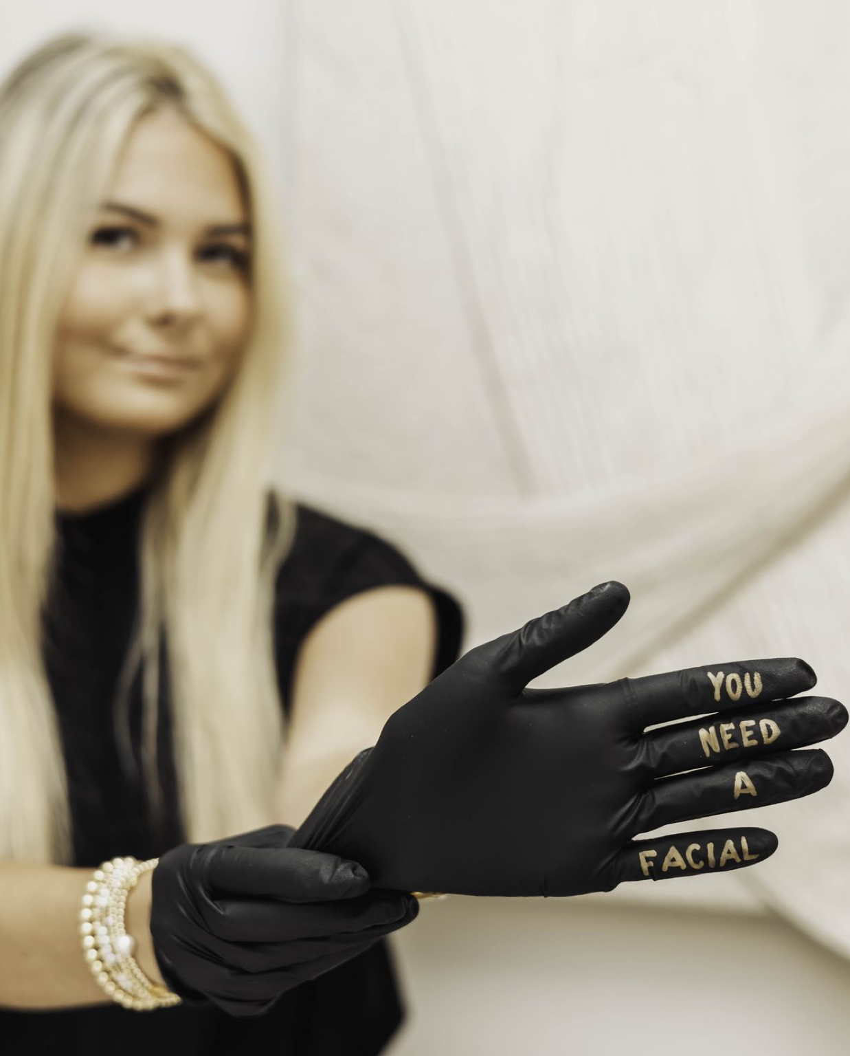 Blonde woman in black top wearing black gloves with gold text reading YOU NEED A FACIAL, promoting skincare services.