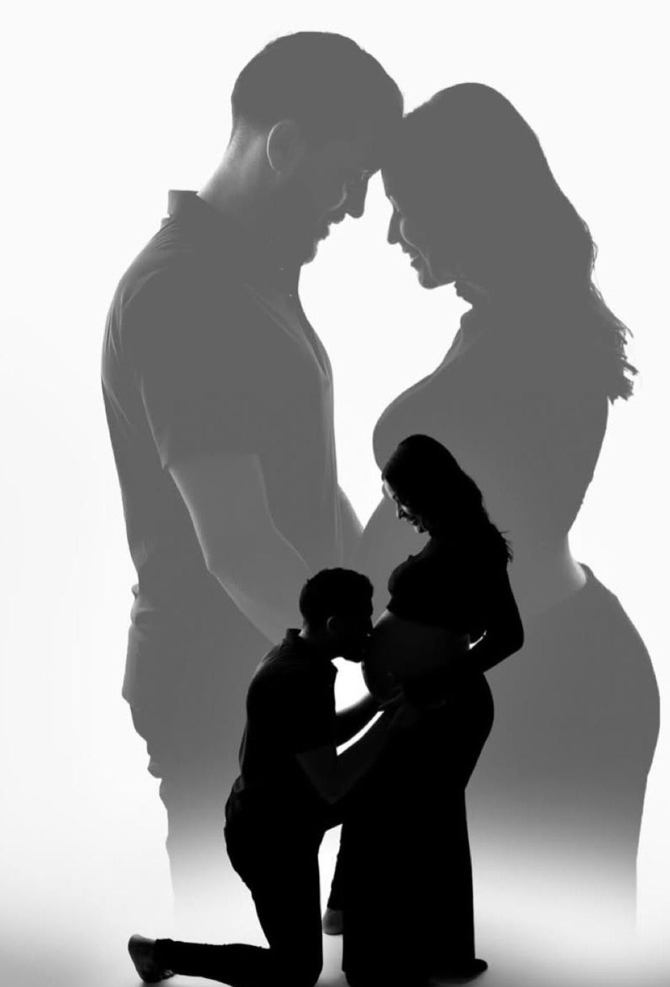 Black and white silhouette of expecting couple, man kneeling kissing pregnant belly, romantic maternity photo.