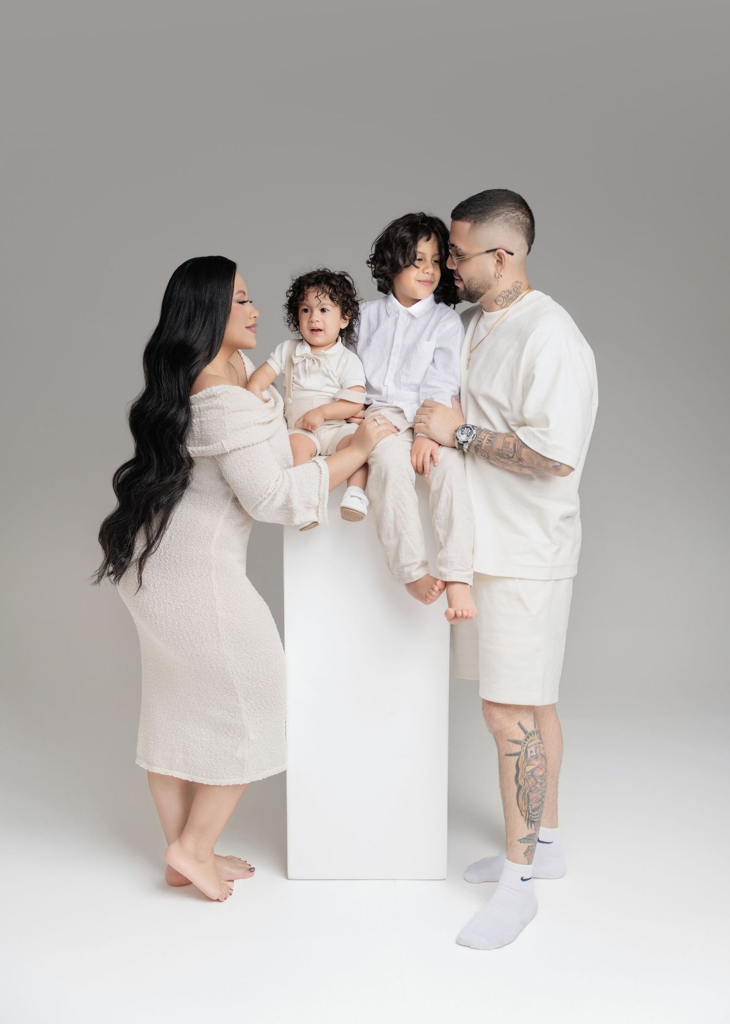 A family of four in matching cream outfits poses together in a studio, with two young children sitting on a white block.