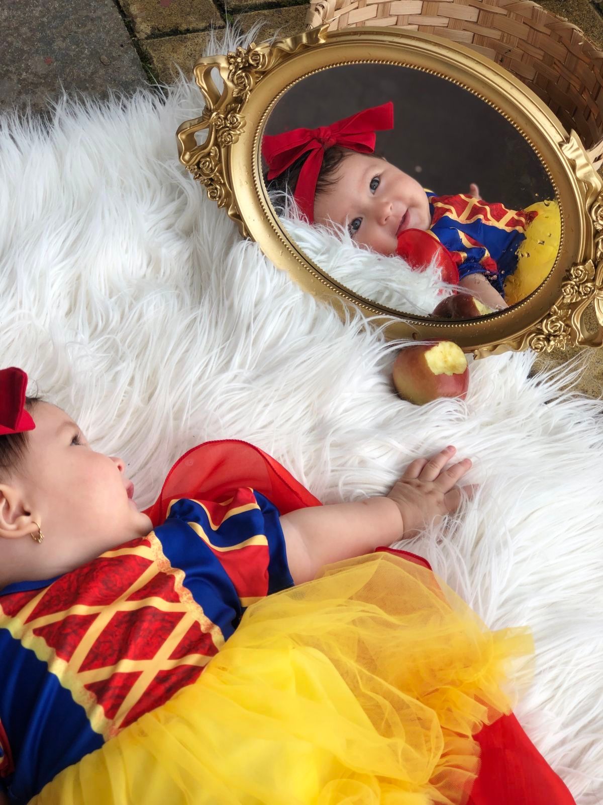 Baby dressed as Snow White lying on white fur rug beside a gold ornate mirror reflecting her smiling face.