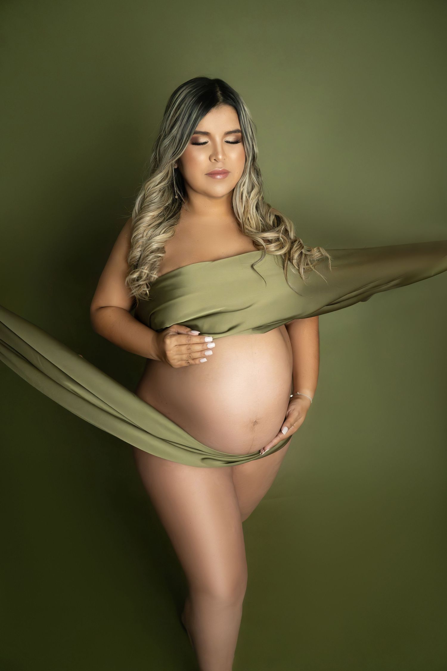 Pregnant woman draped in olive green fabric poses against matching background, cradling her bare baby bump.