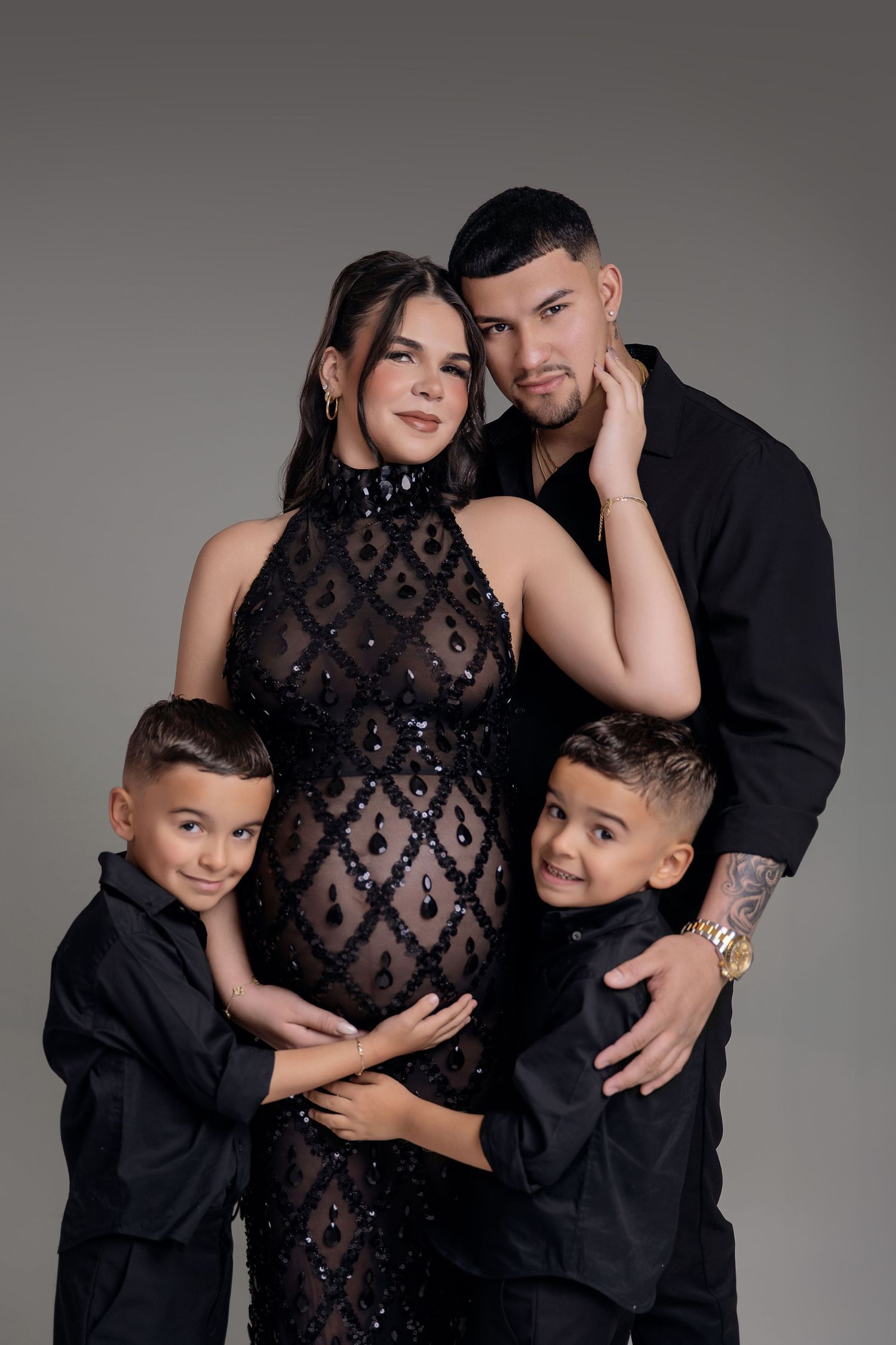 Pregnant woman in black sequin dress poses with husband and two young sons in matching black outfits for maternity photo.