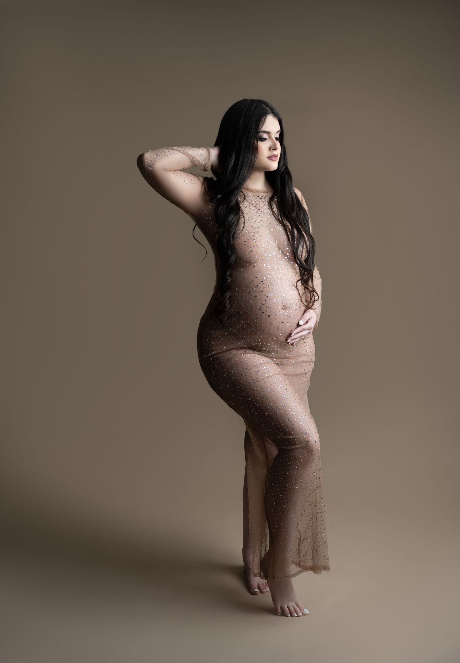 Pregnant woman in sheer lace nude gown poses gracefully, cradling baby bump in professional maternity studio portrait.