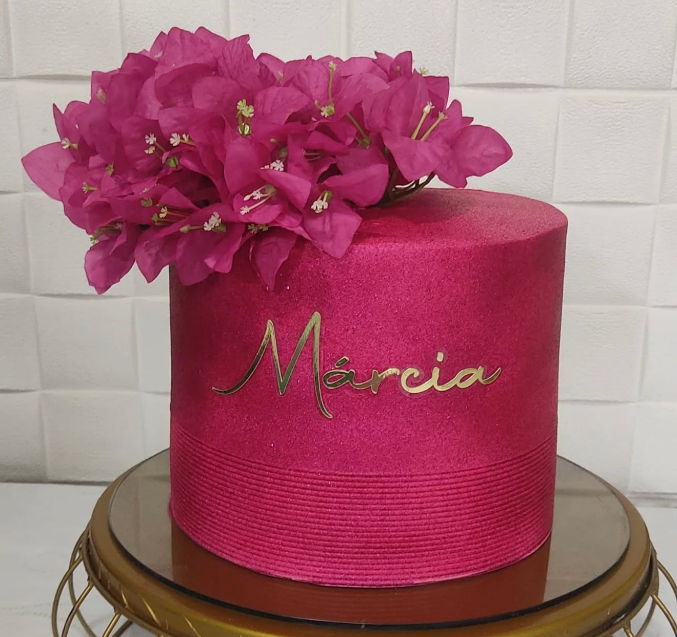 Hot pink glittery cake topped with fuchsia bougainvillea flowers, decorated with gold script name Marcia on a gold cake stand.
