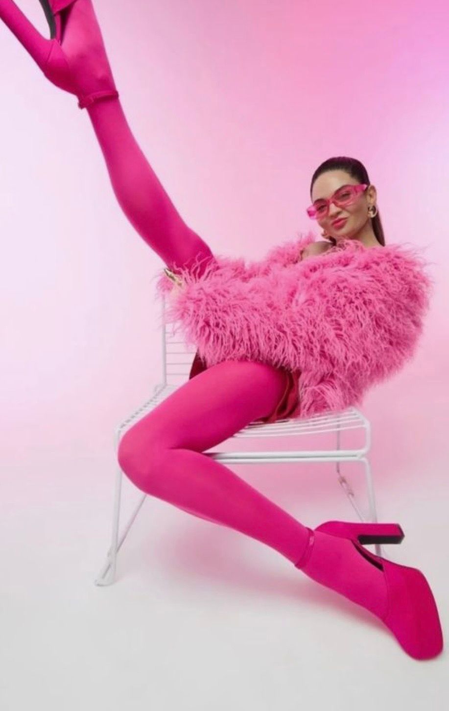 Woman in pink feathered jacket, hot pink tights and heels poses on white chair against pink background.
