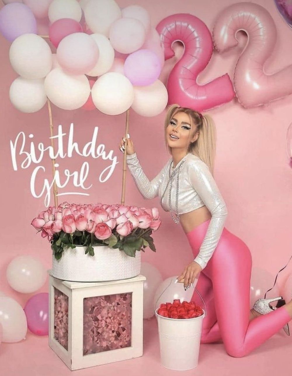 A blonde woman poses in pink outfit beside floral arrangement and balloons for a 22nd birthday celebration.