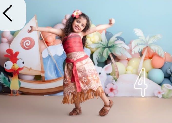Young girl in Moana costume poses dancing at a tropical themed 4th birthday party with colorful balloon decorations.