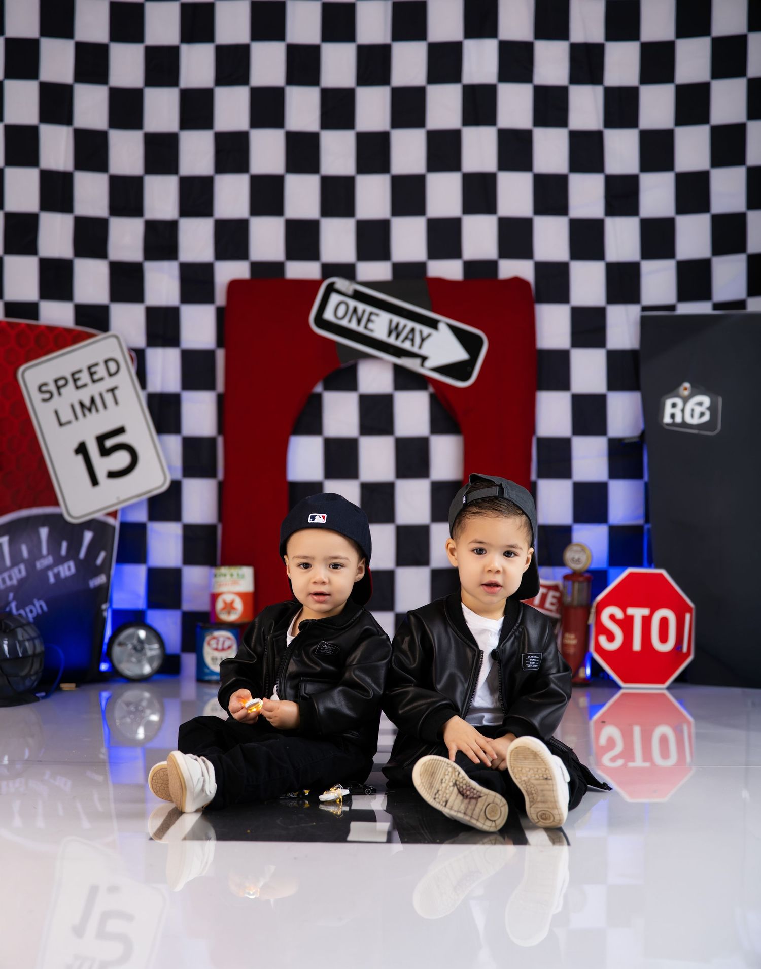 Two young children in black leather jackets sit before a checkered racing backdrop with street signs at a themed photo shoot.