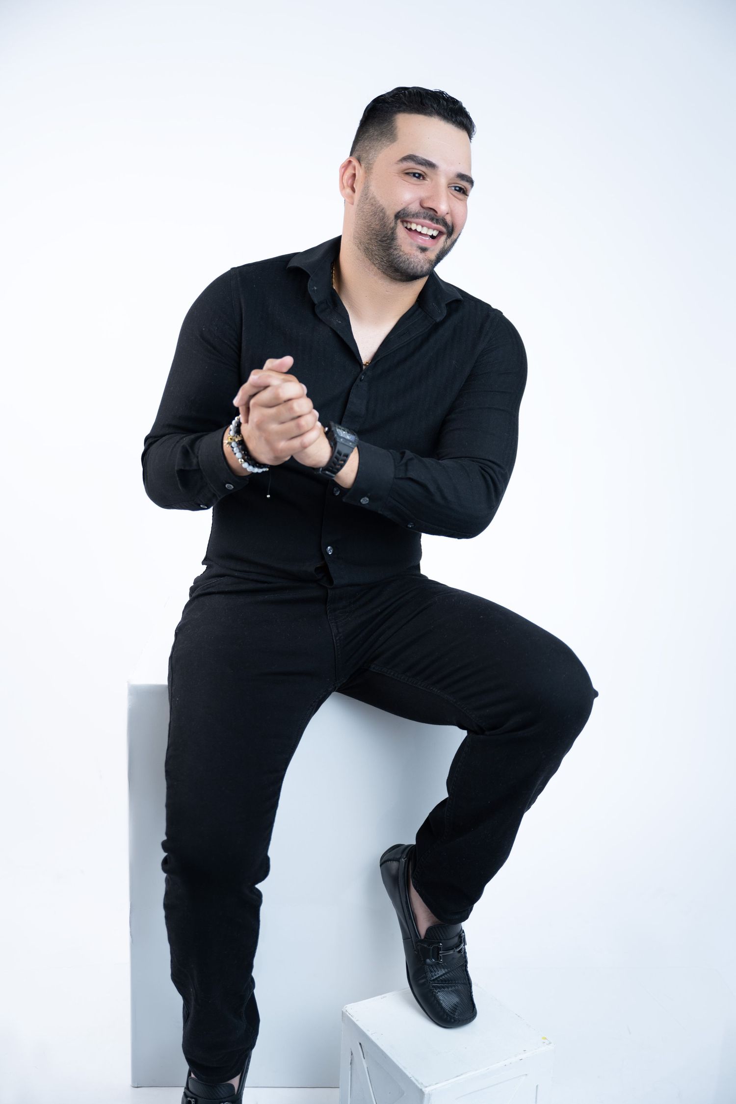 Professional in all-black attire seated on white cube with clasped hands.