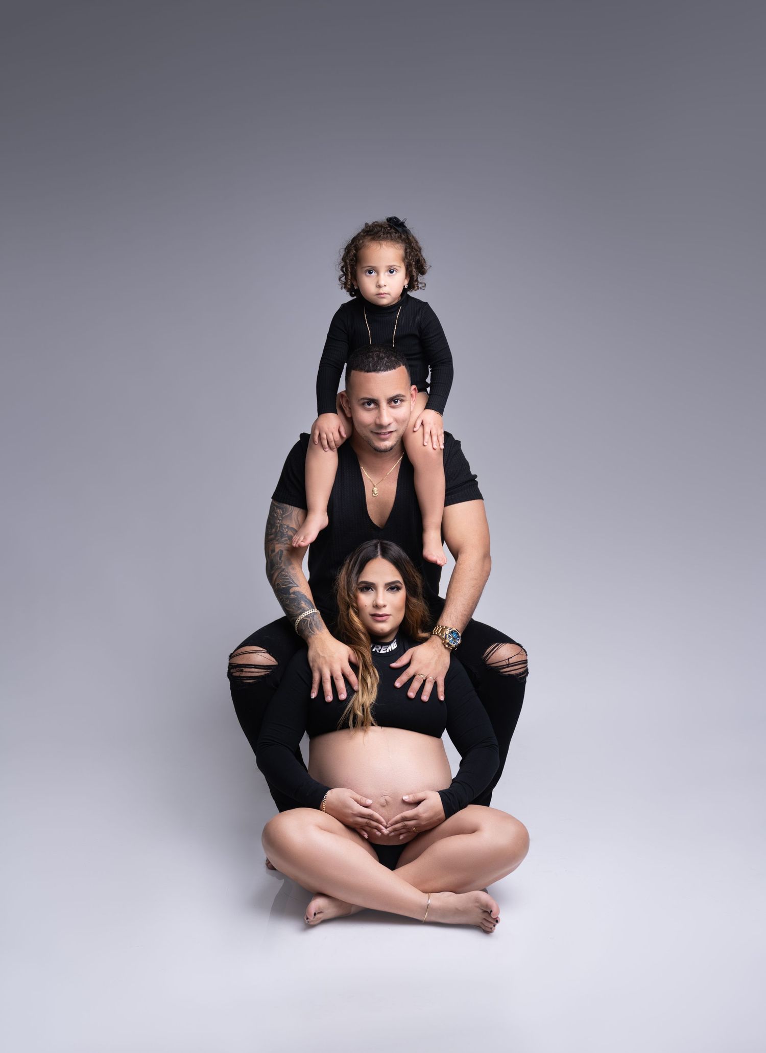 A family maternity portrait with three people sitting on grey backdrop.
