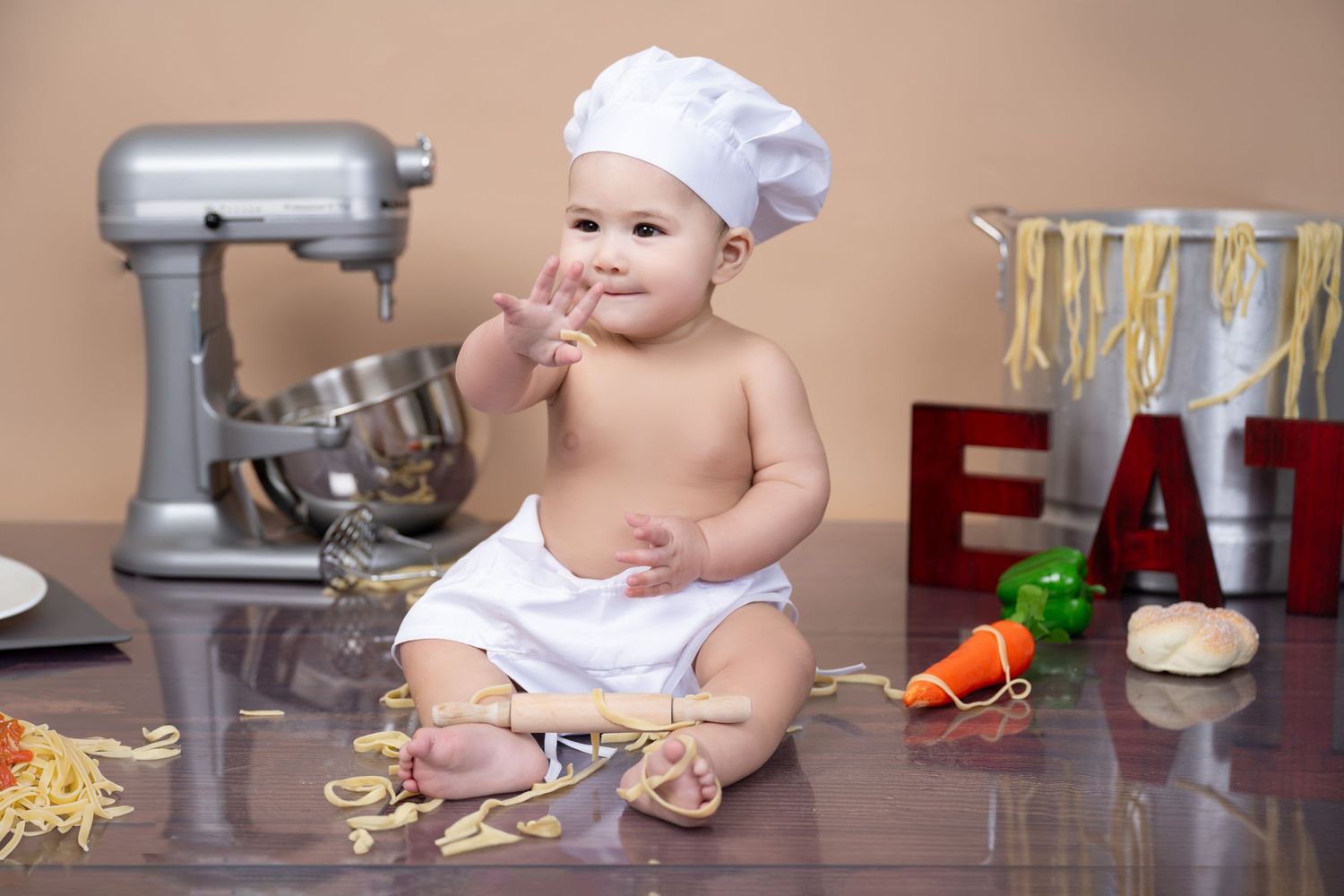 Small child in chef hat sits on counter with stand mixer and pasta ingredients in kitchen setting.