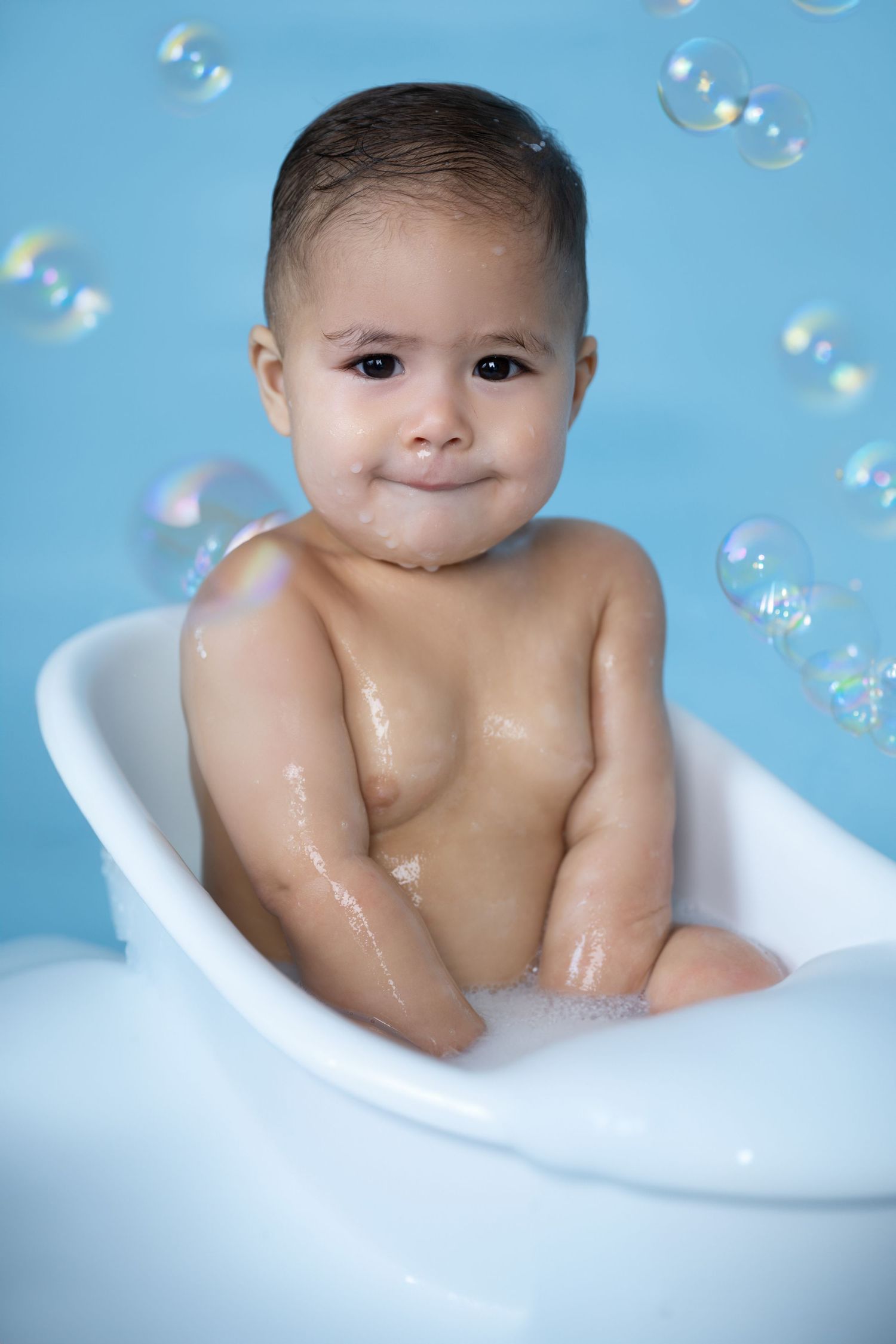 Child sits in white bathtub with soap bubbles floating against blue background.