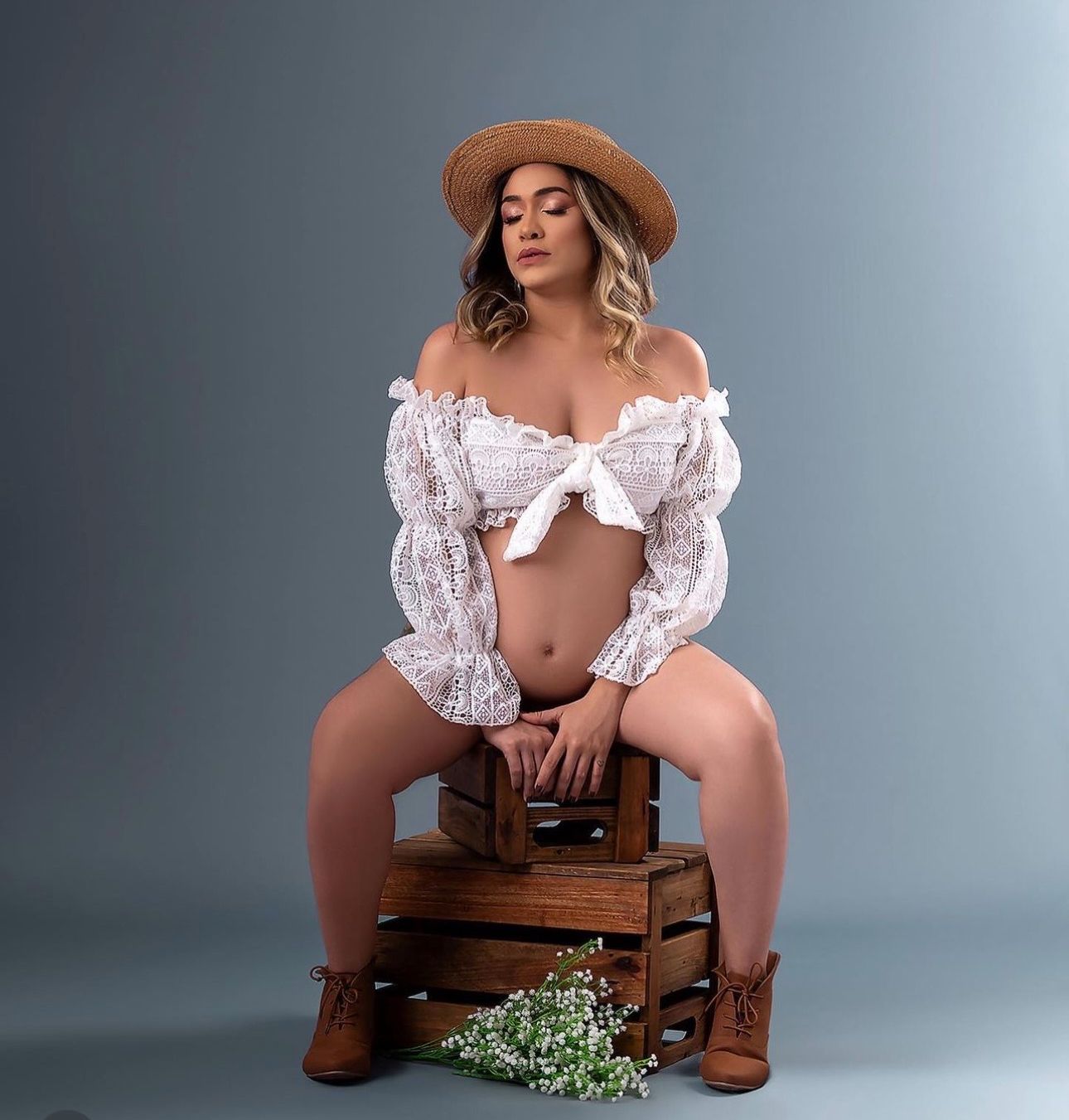 Woman in white crop top and hat sitting on wooden crate with flowers in a studio setting.