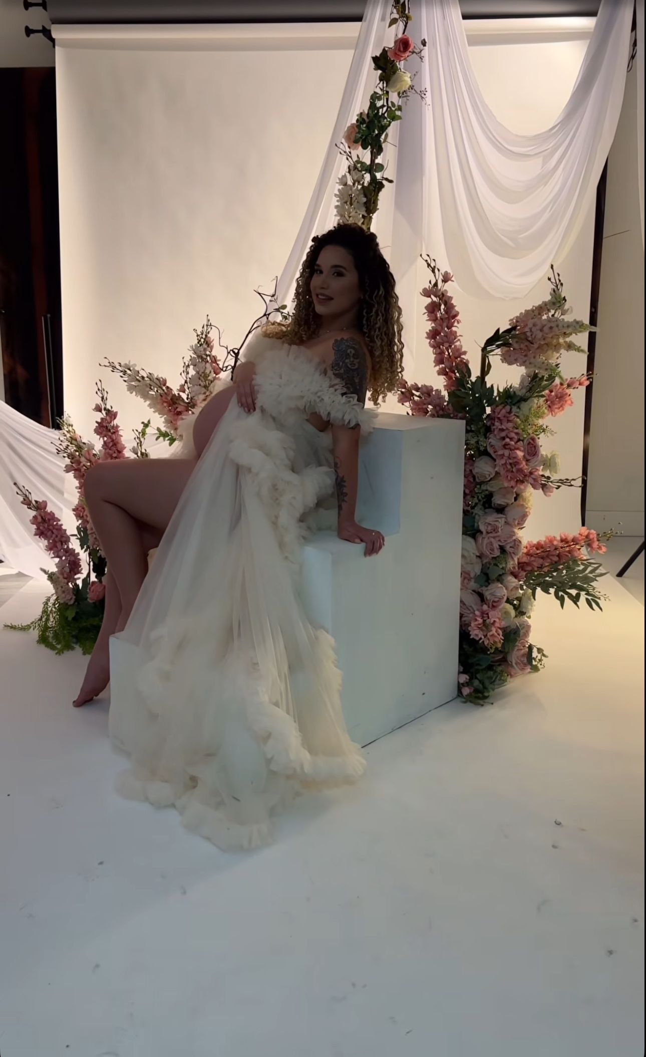 Woman in white ruffled off-shoulder gown poses in floral studio with pink blooms and draped white backdrop.