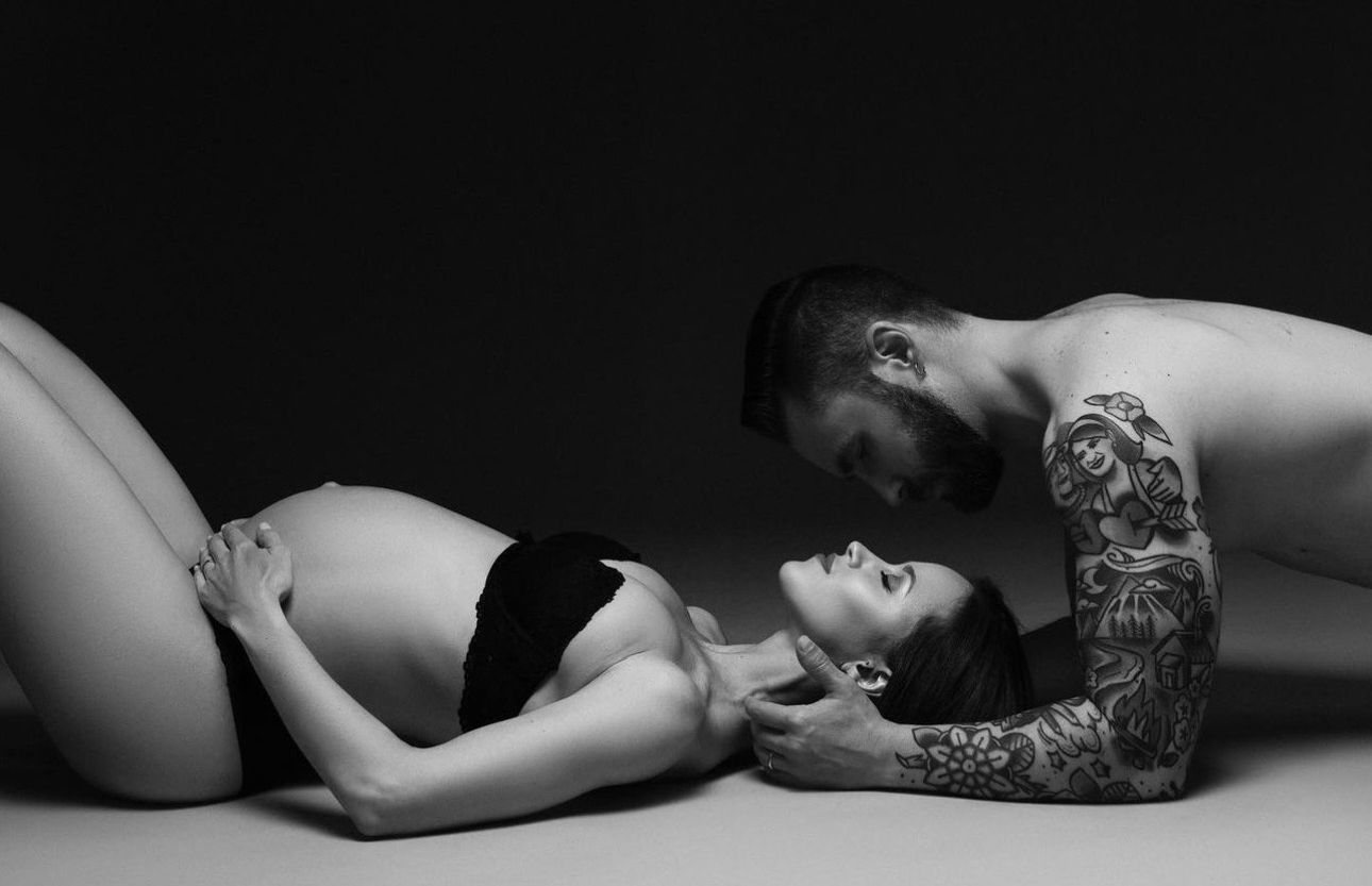 Black and white maternity photo of pregnant woman lying down with tattooed partner leaning over her tenderly.