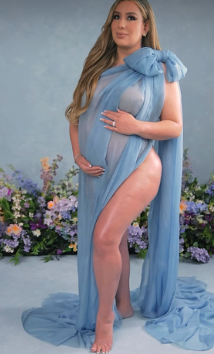 A maternity photoshoot featuring a flowing light blue sheer fabric drape against a floral backdrop.